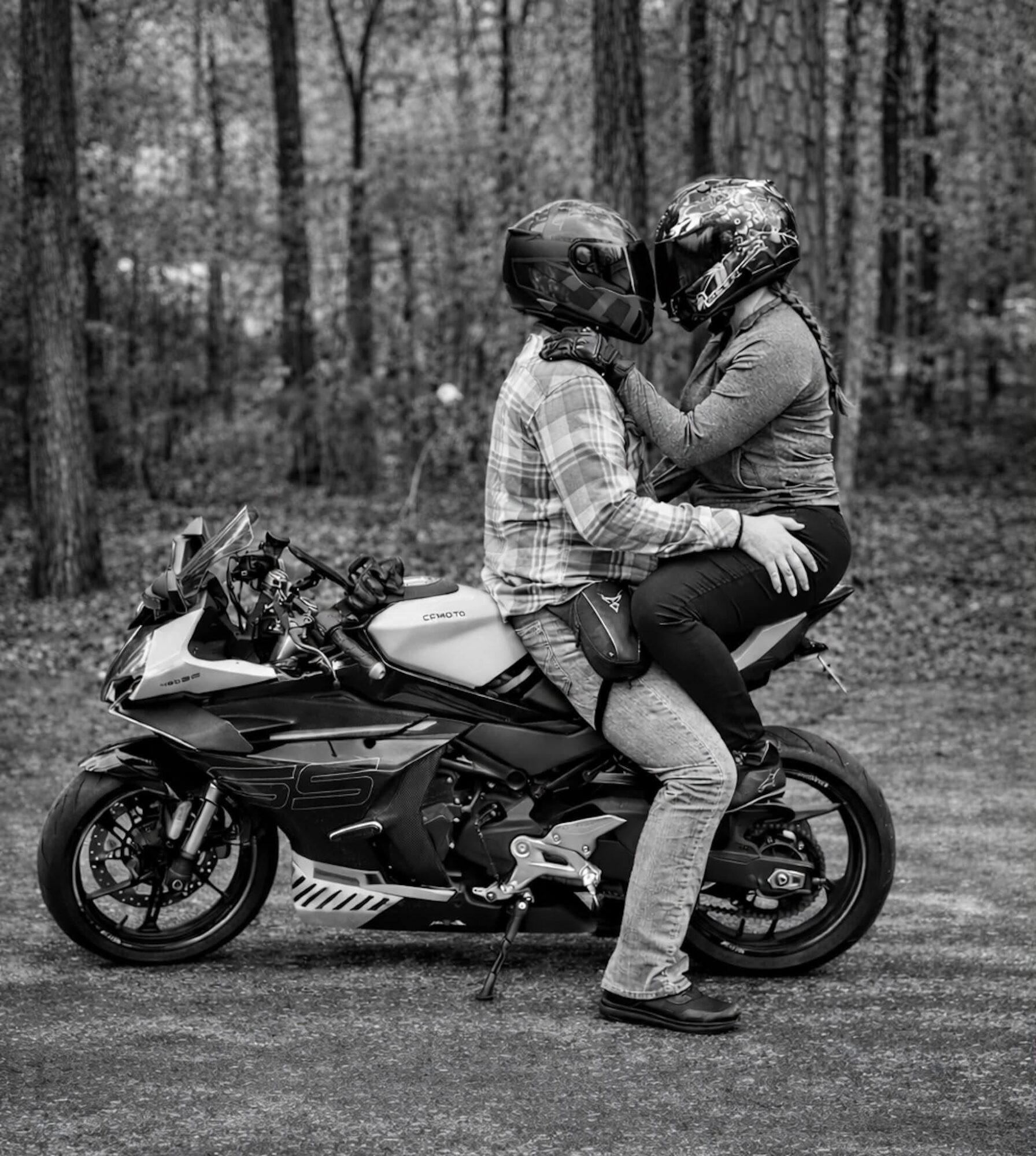 Two people sitting on a motorcycle in a forest, wearing helmets and casual clothing, facing each other closely.