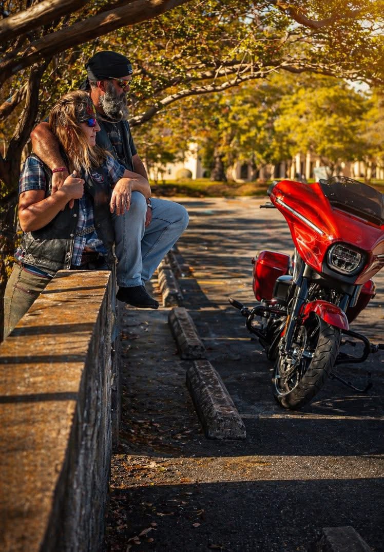 Two people, one with a beard and glasses, stand near a red motorcycle parked on the street under trees with autumn leaves.