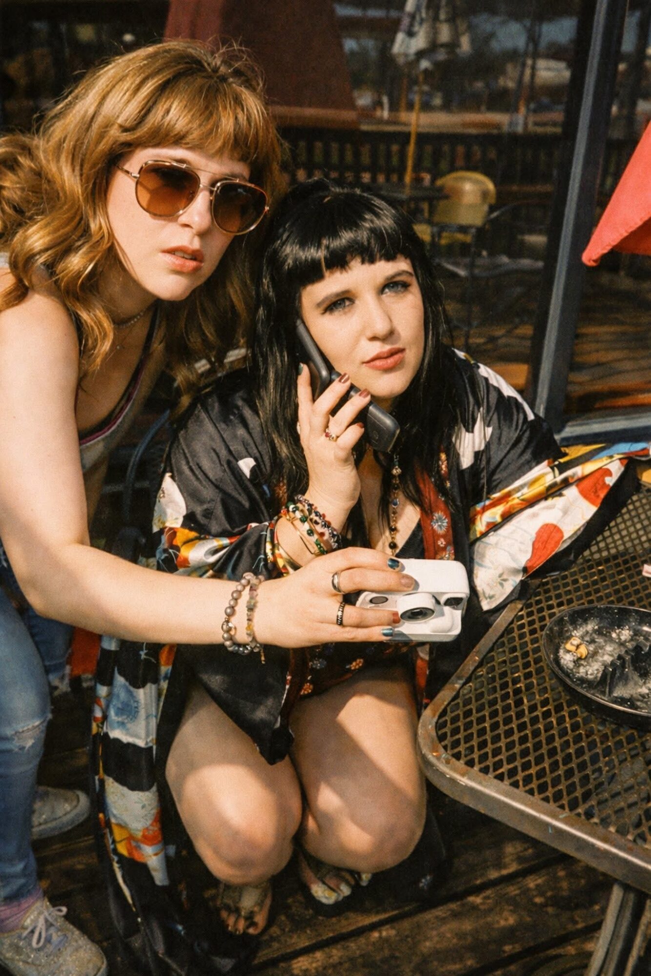 Two women sitting at an outdoor table, one with sunglasses and wavy hair, the other with straight black hair, holding a phone.