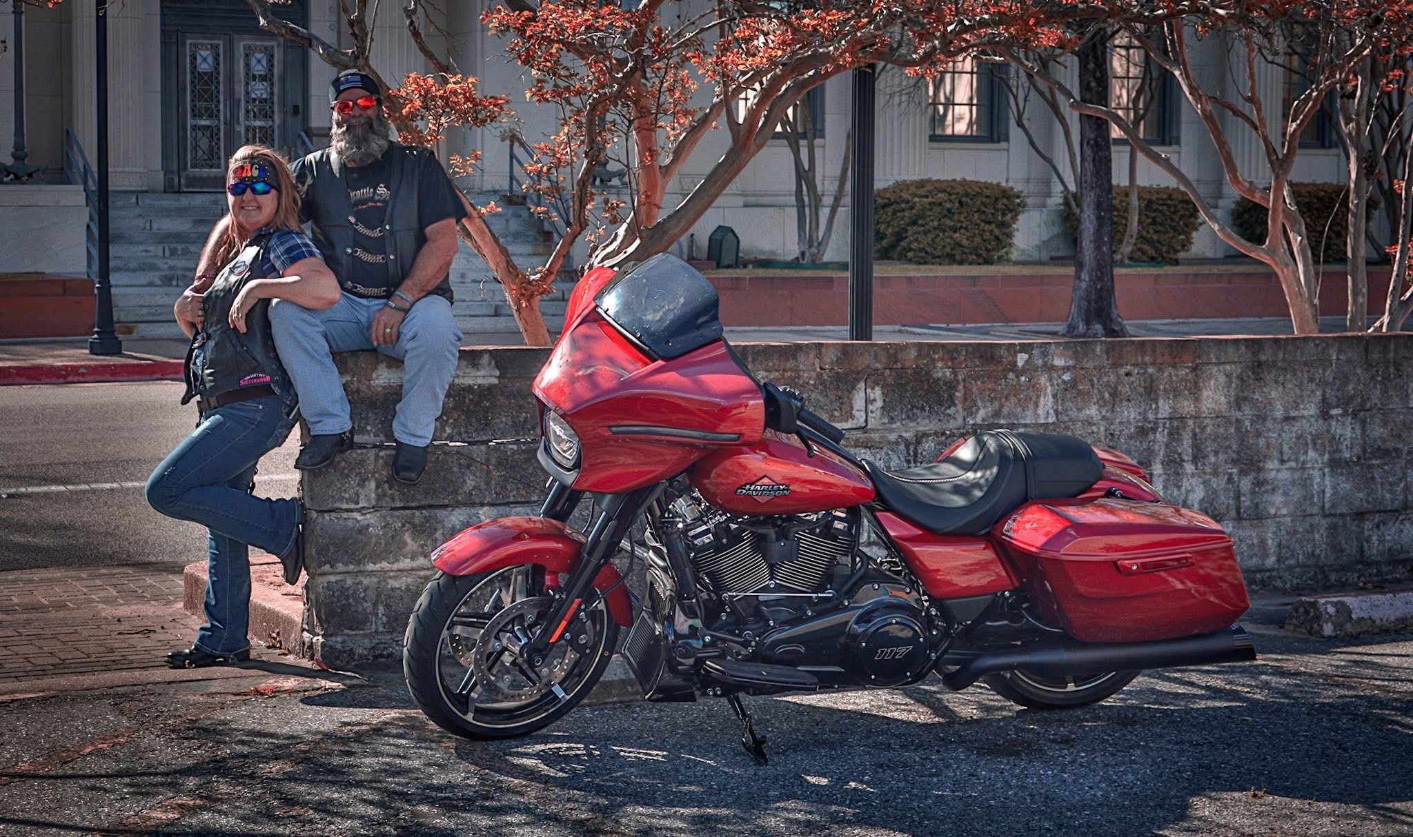 Two people sit on a bench behind a red motorcycle parked on the street, with trees and buildings in the background.