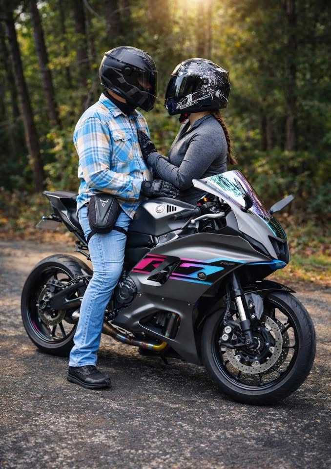 Two people wearing helmets stand close together beside a black motorcycle on a forested road.