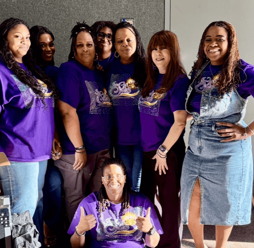 Group of seven women, one sitting in front, all wearing purple shirts, smiling, indoors against a gray wall.
