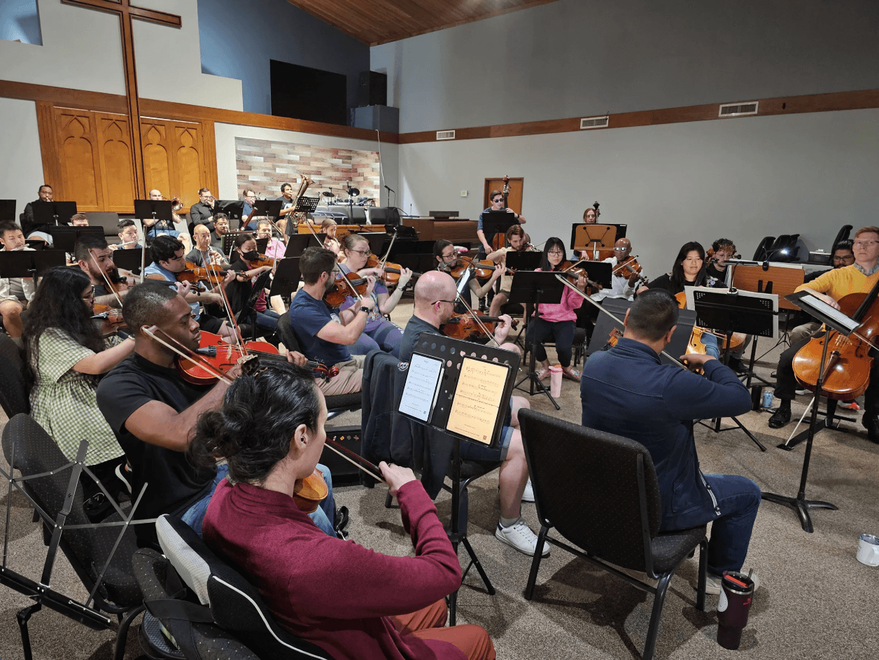 Orchestra performing in a church or concert hall with musicians playing violins, cellos, and other instruments, audience in background.