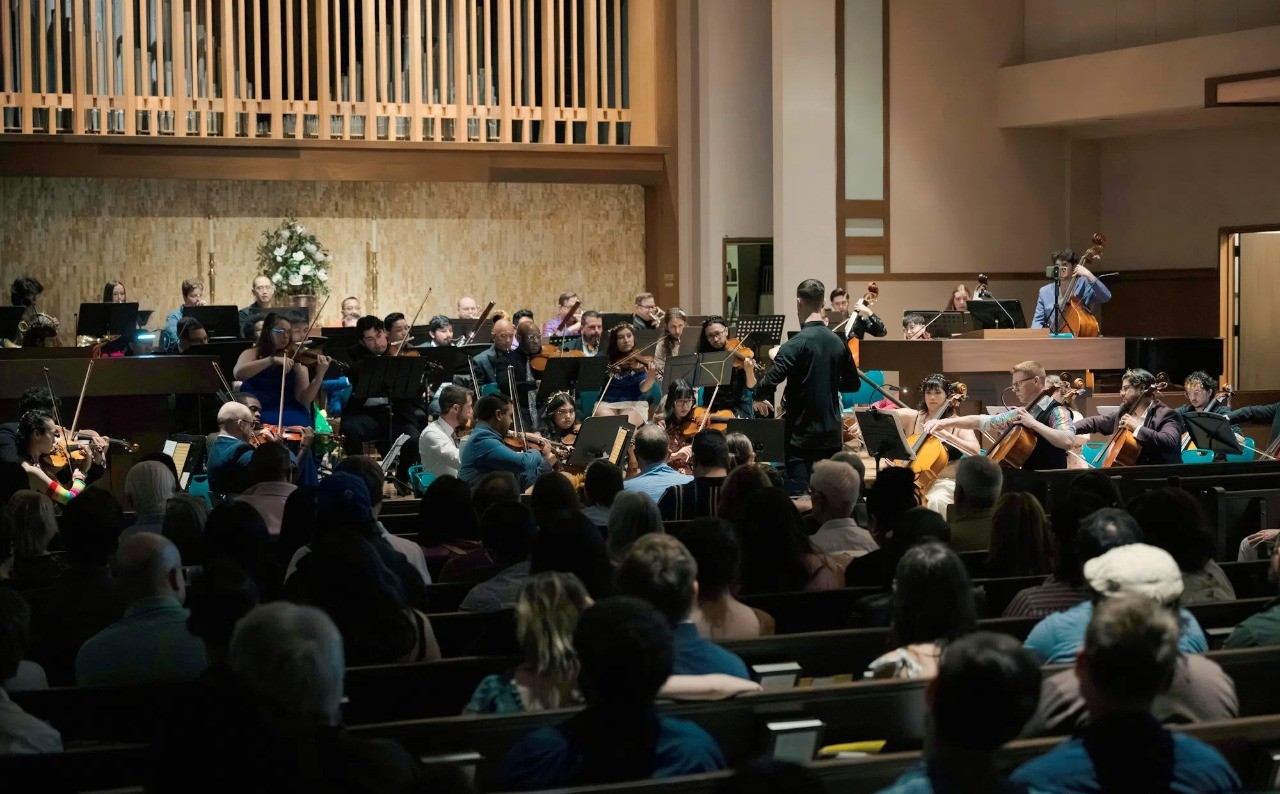 Orchestra performing in a concert hall with audience watching, conductor leading, and musicians playing instruments.