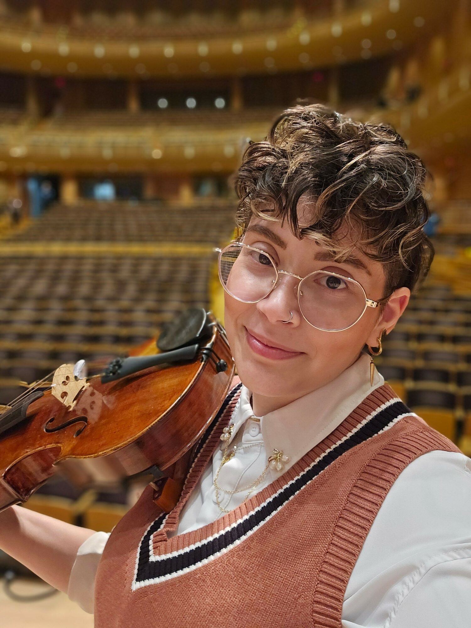 Person with curly hair and glasses holding a violin, smiling, in a room with shelves and warm lighting.