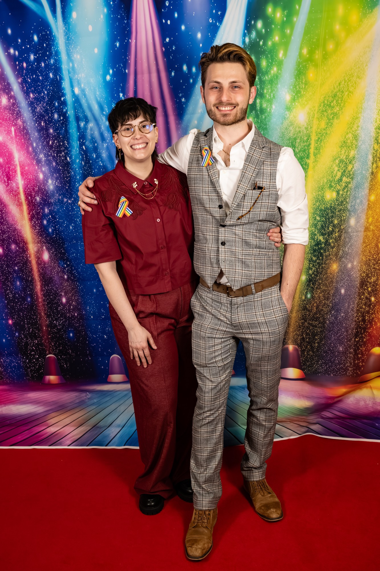 A smiling woman in a maroon uniform and a man in a gray plaid suit stand together on a red carpet, colorful backdrop behind them.
