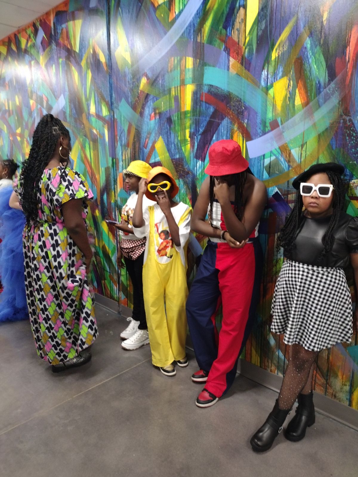 Group of children and a woman in front of a colorful abstract mural, some wearing sunglasses and hats, standing and talking.