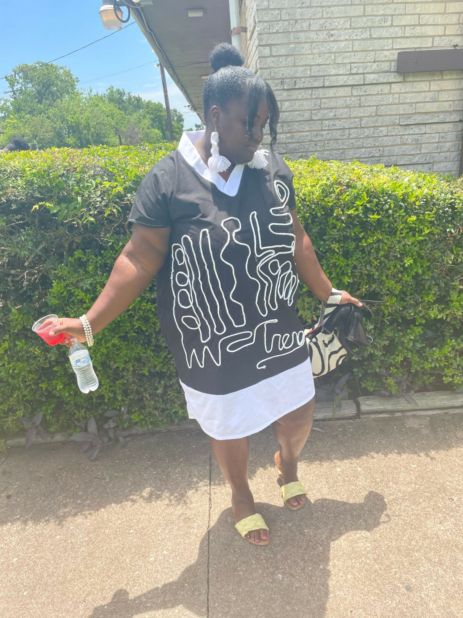 Person walking outdoors holding a water bottle and a red object, wearing a black and white patterned dress and sandals.