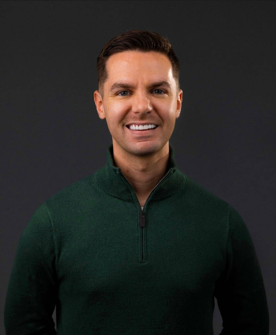 Smiling man with short dark hair wearing a green zip-up sweater against a dark background.