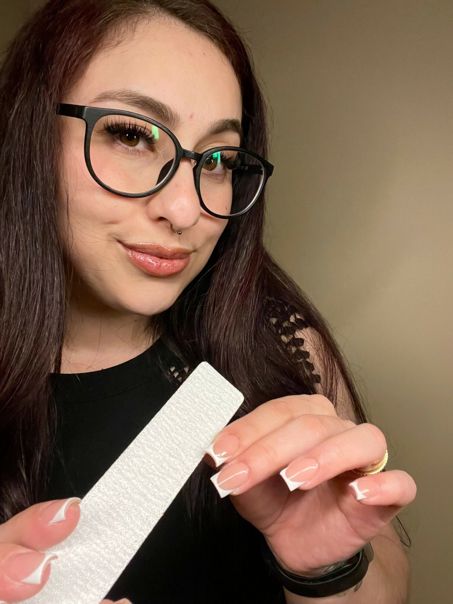 Young woman with glasses holding a nail file, smiling, with long dark hair and a black top, against a plain background.
