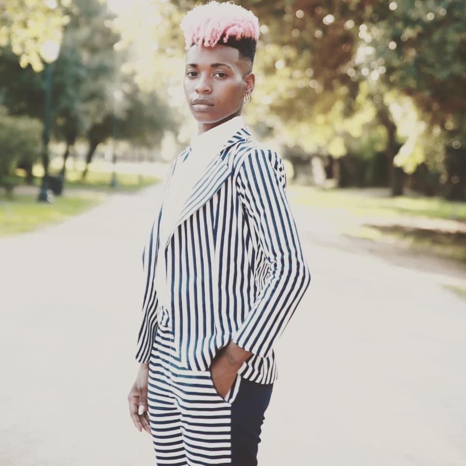 Young person with short pink hair wearing a striped blazer and pants, standing outdoors on a sunny day.