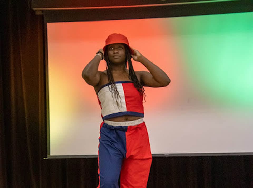 A person stands in front of a colorful background, wearing a red hat, a sleeveless top, and red and blue pants.