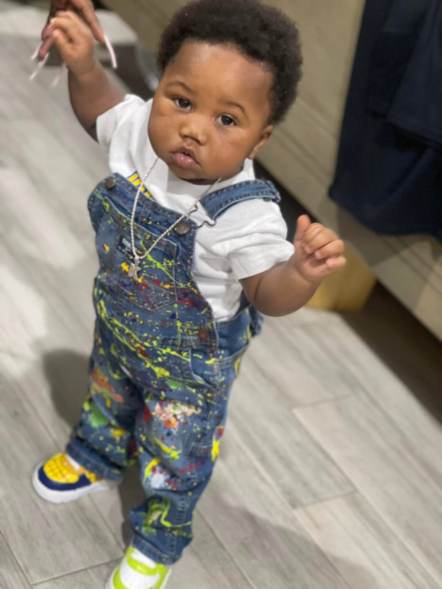 Young child with curly hair wearing overalls and sneakers, standing on a wooden floor, looking at the camera.