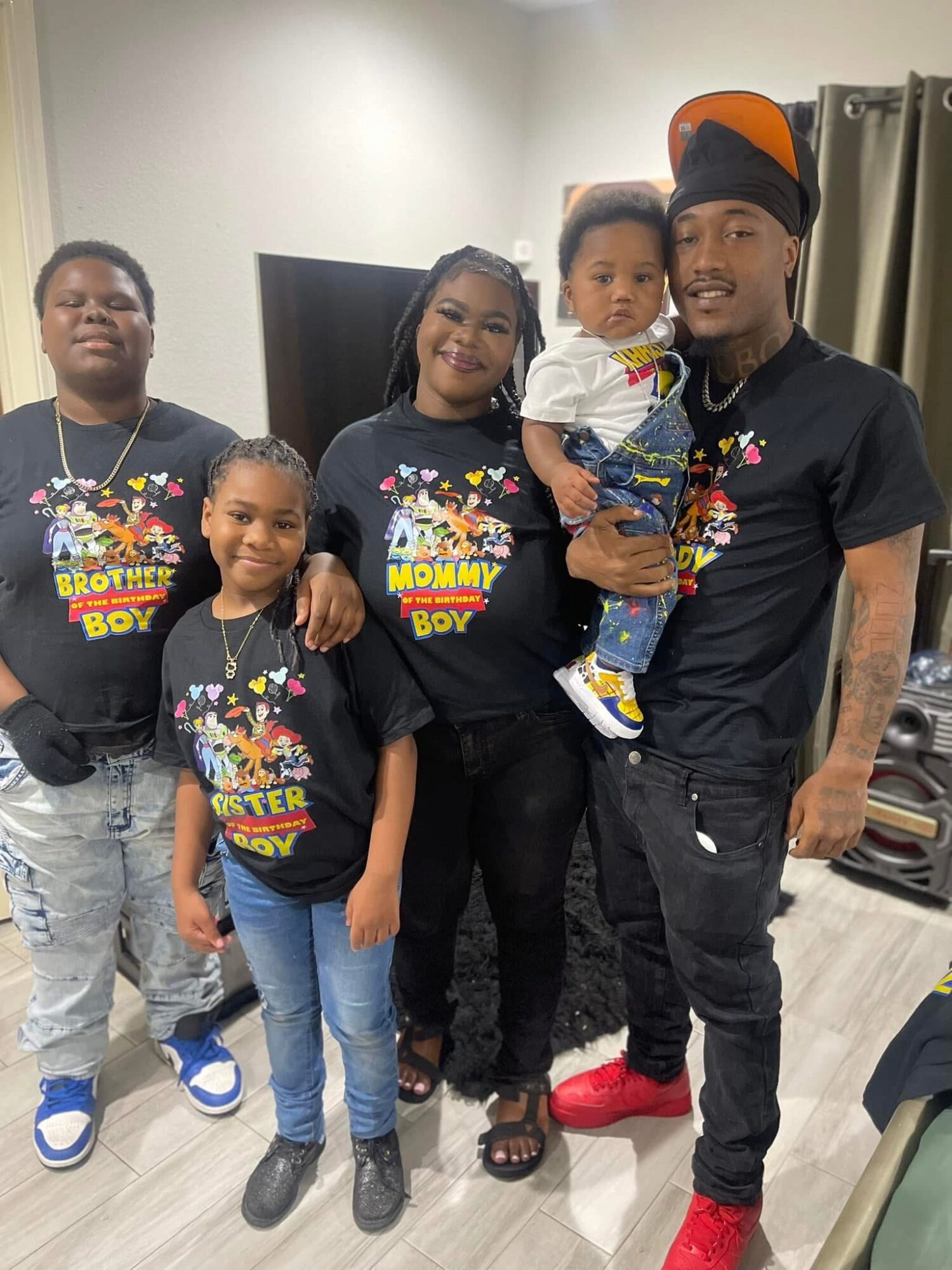 Group of five people, including children and adults, standing indoors, smiling, wearing matching black shirts with colorful text, in a living room.