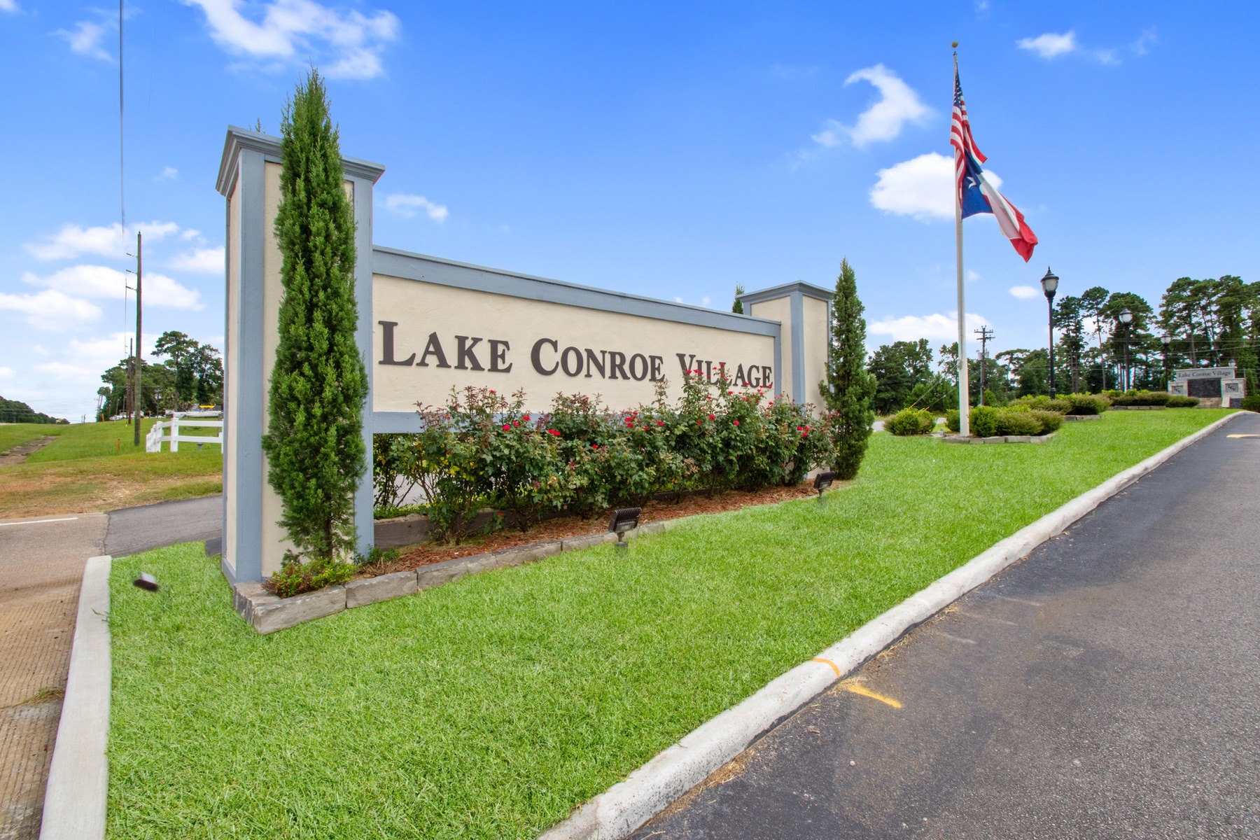 Sign reading 'Lake Conroe Village' with greenery and flags, under a blue sky.