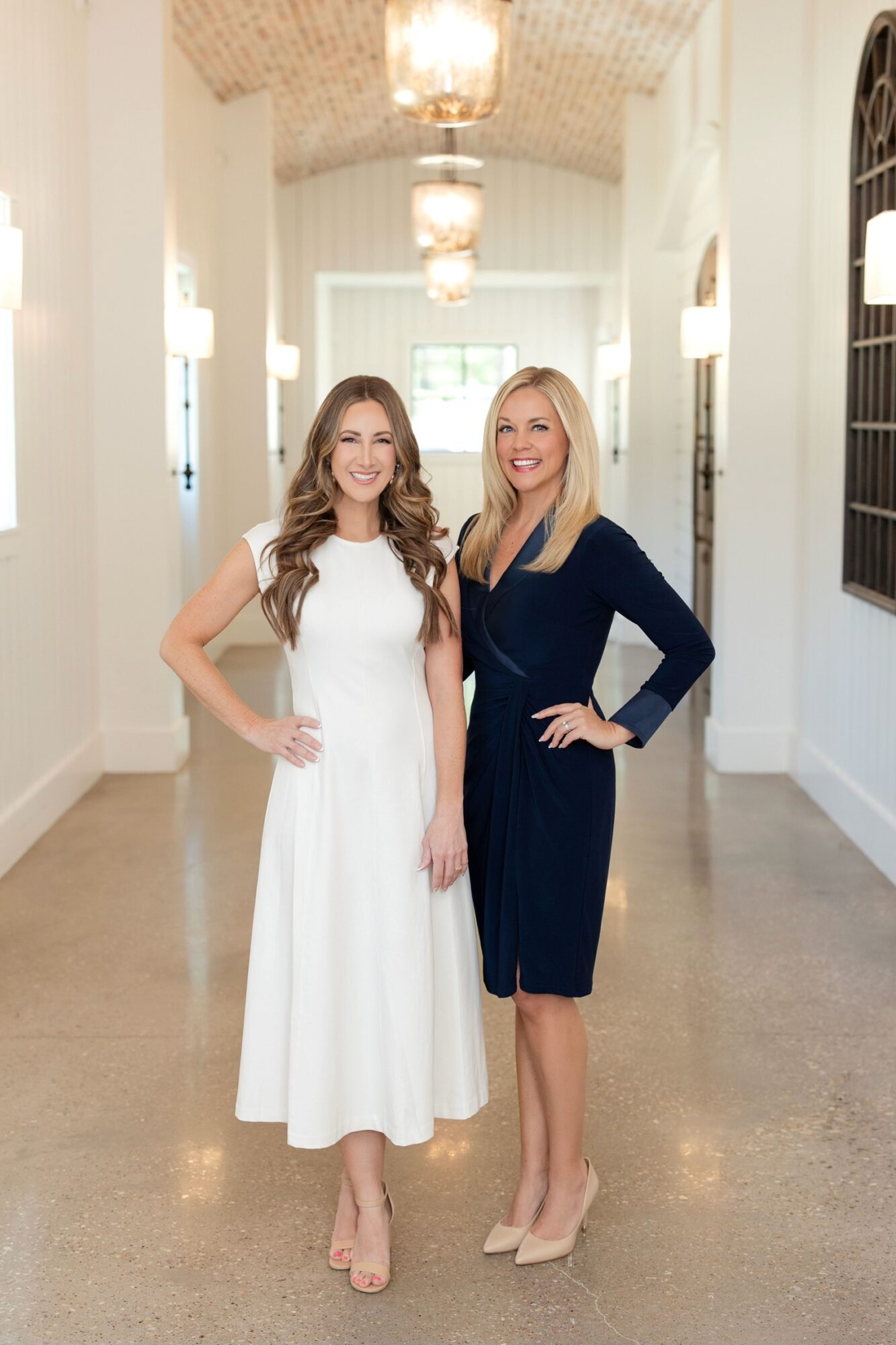 Two women stand in a bright, elegant hallway with chandeliers, smiling at the camera, one in a white dress, the other in a navy dress.