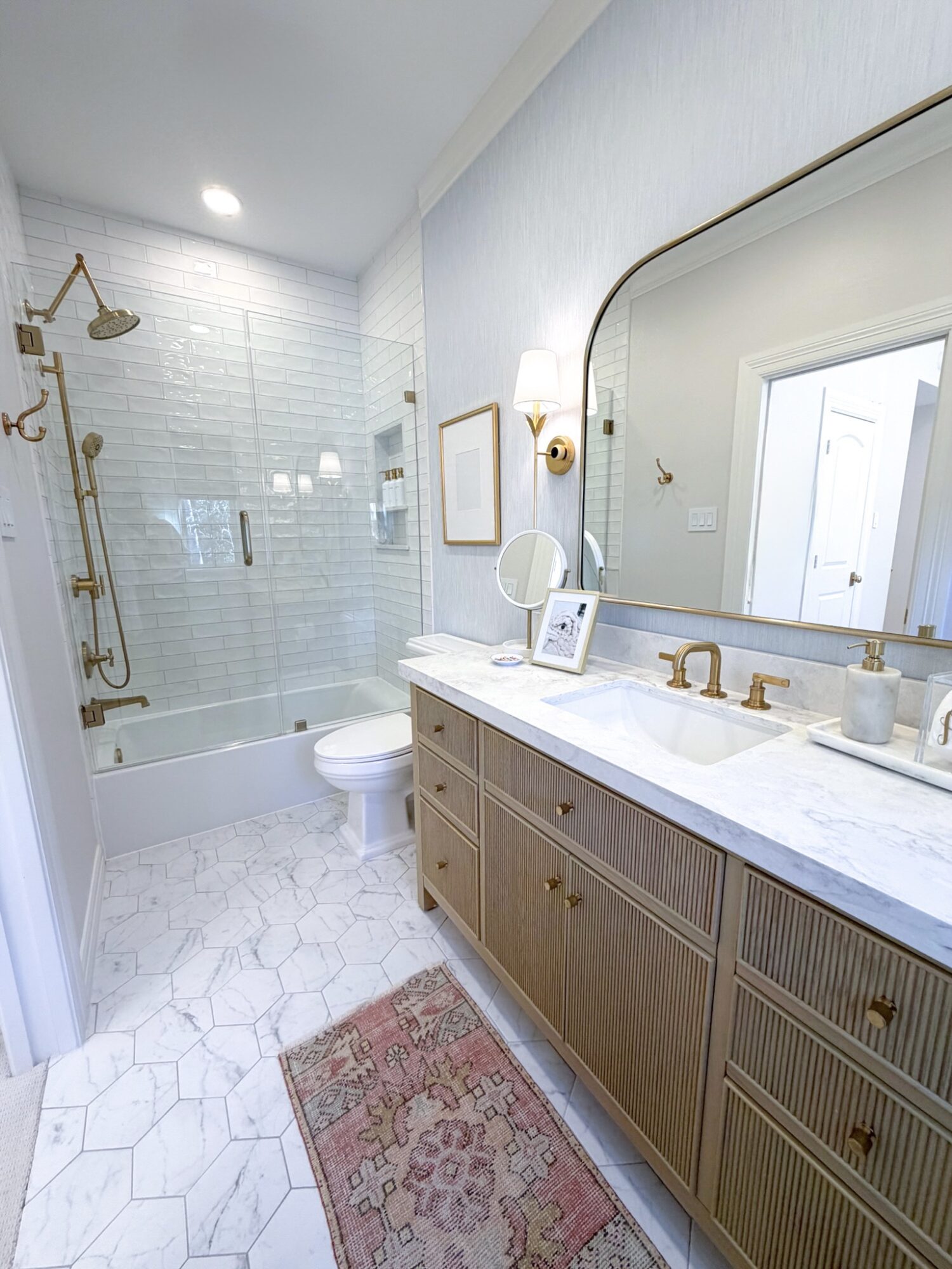 Bathroom with a large mirror, vanity with gold fixtures, and a shower with glass door, white walls, and hexagonal tile floor.
