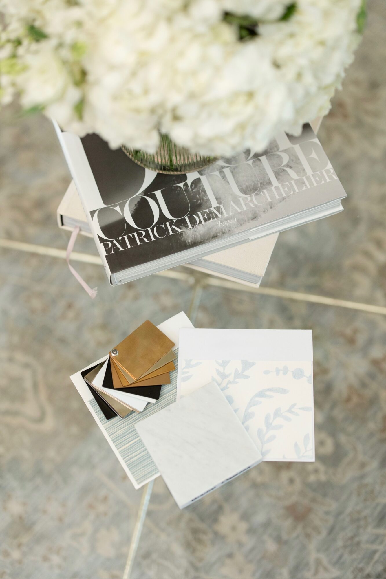 A glass table with a bouquet of white flowers, a magazine, and color swatches on a tiled floor.