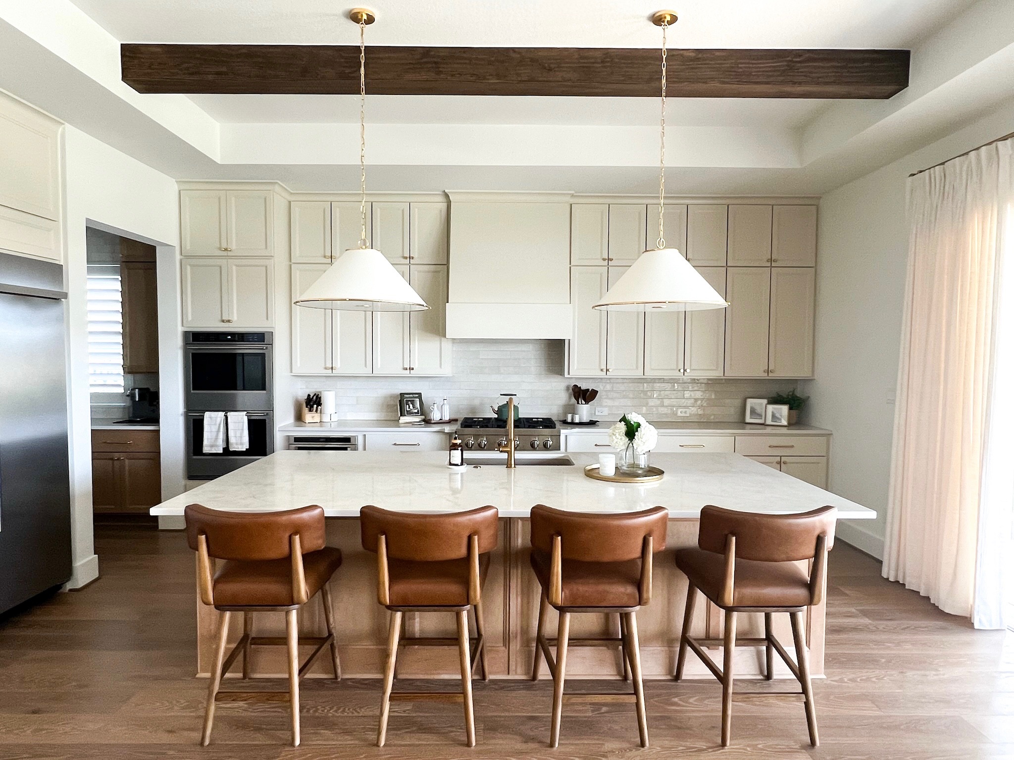 Kitchen with white cabinets, a large island, five brown chairs, and pendant lights, with a window and curtains on the right.