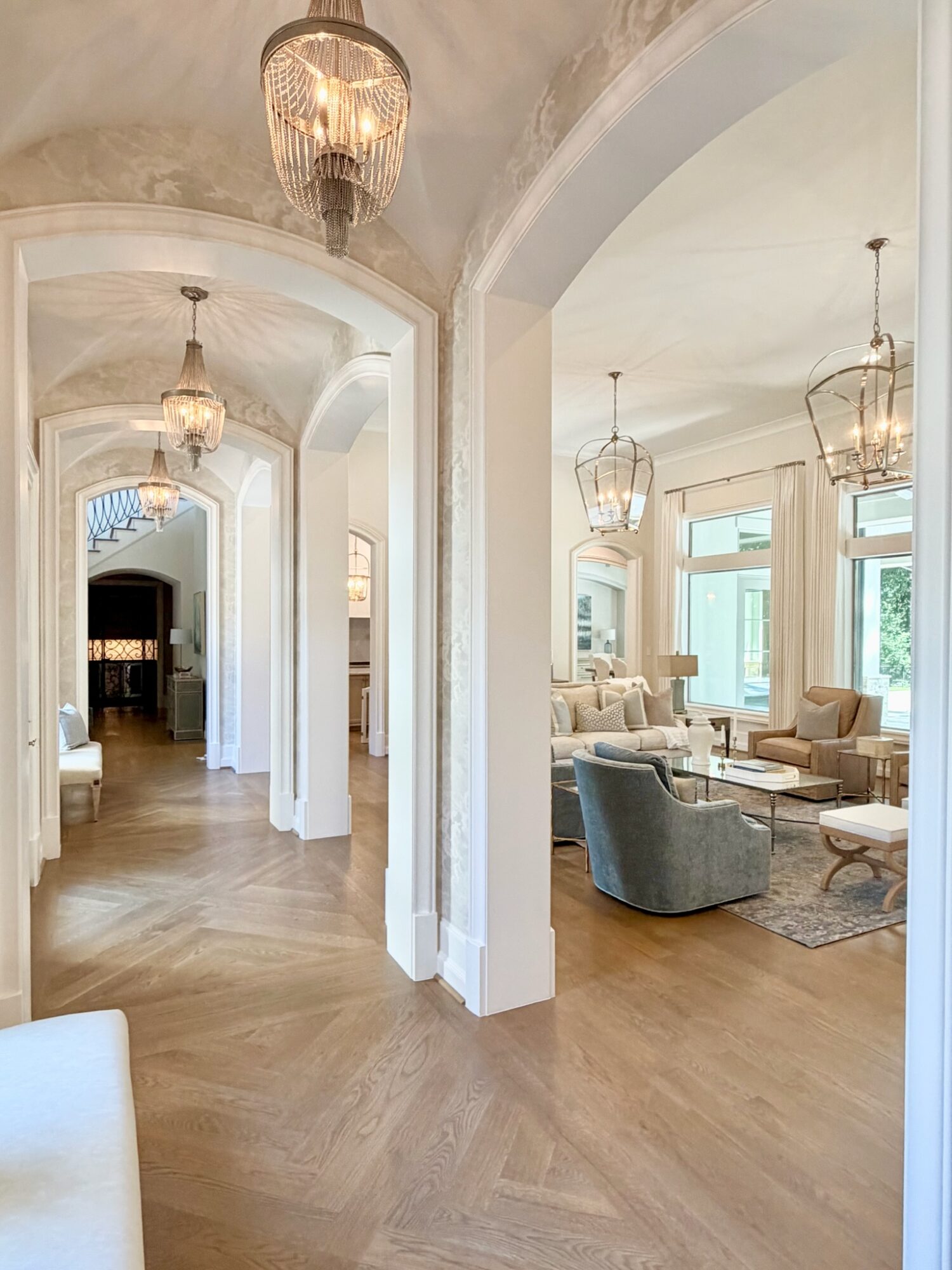 Elegant hallway with chandeliers, leading to a bright living room with large windows and comfortable seating.