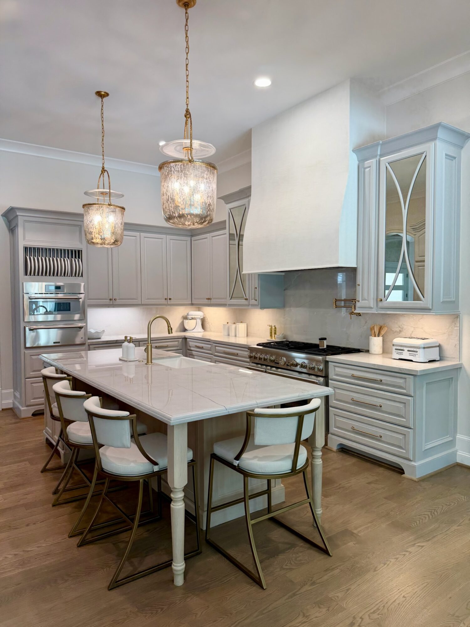 Modern kitchen with light gray cabinets, white island, and pendant lights, featuring a stove, oven, and glass-front cabinet.