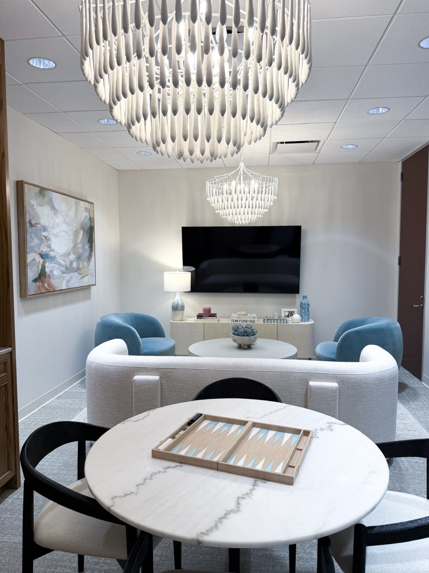Living room with two blue chairs, a white sofa, a wall-mounted TV, and a round marble table with a tray, chandelier, and artwork.