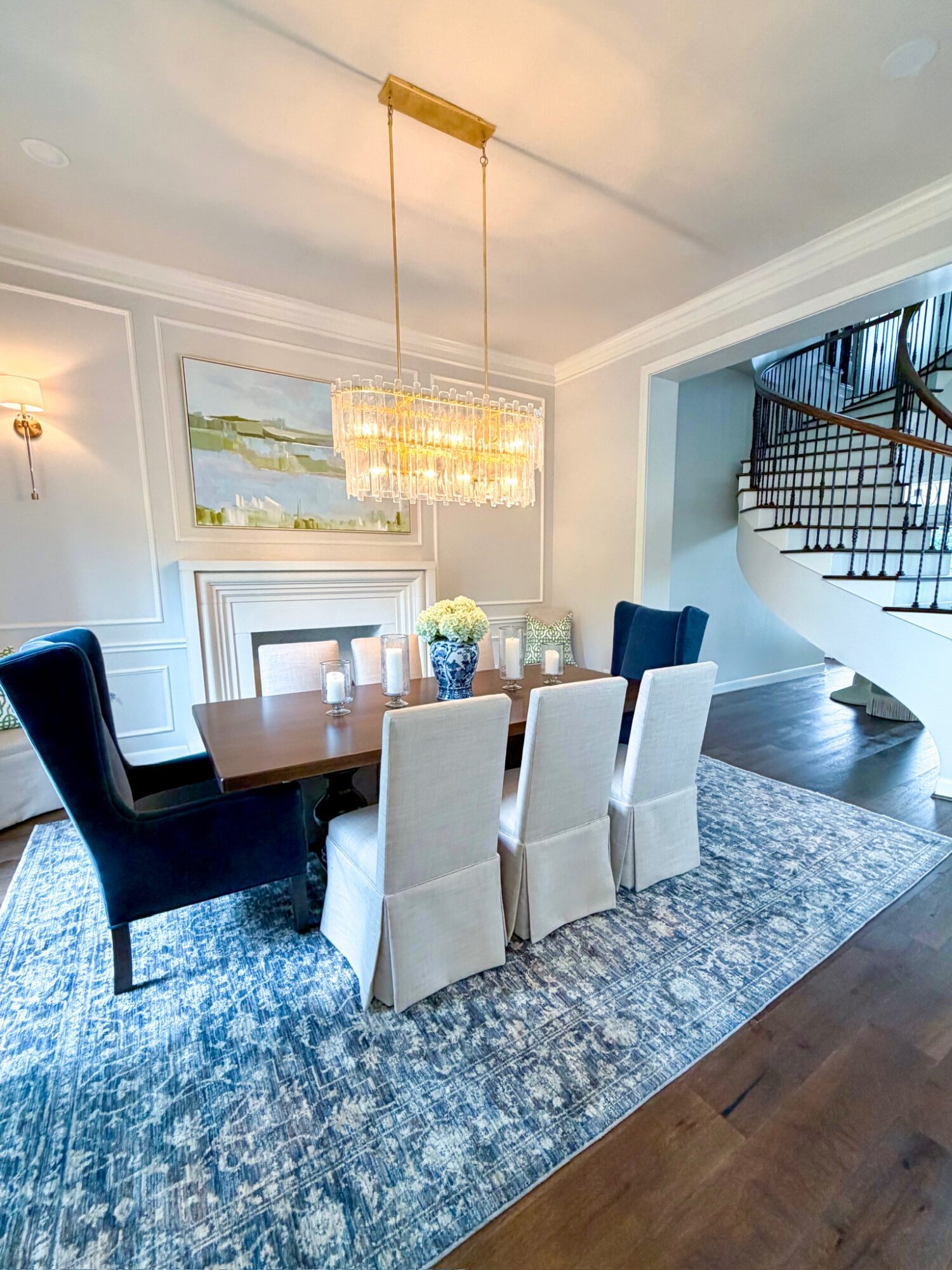 Dining room with a wooden table, six chairs, a chandelier, and a painting on the wall, with a staircase nearby.