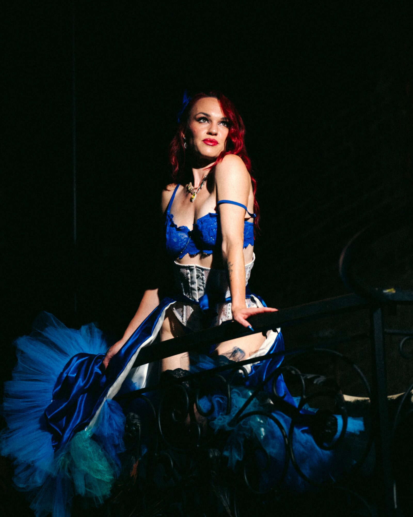 Woman with red hair in blue and black outfit, sitting on a dark stage, looking to the side.