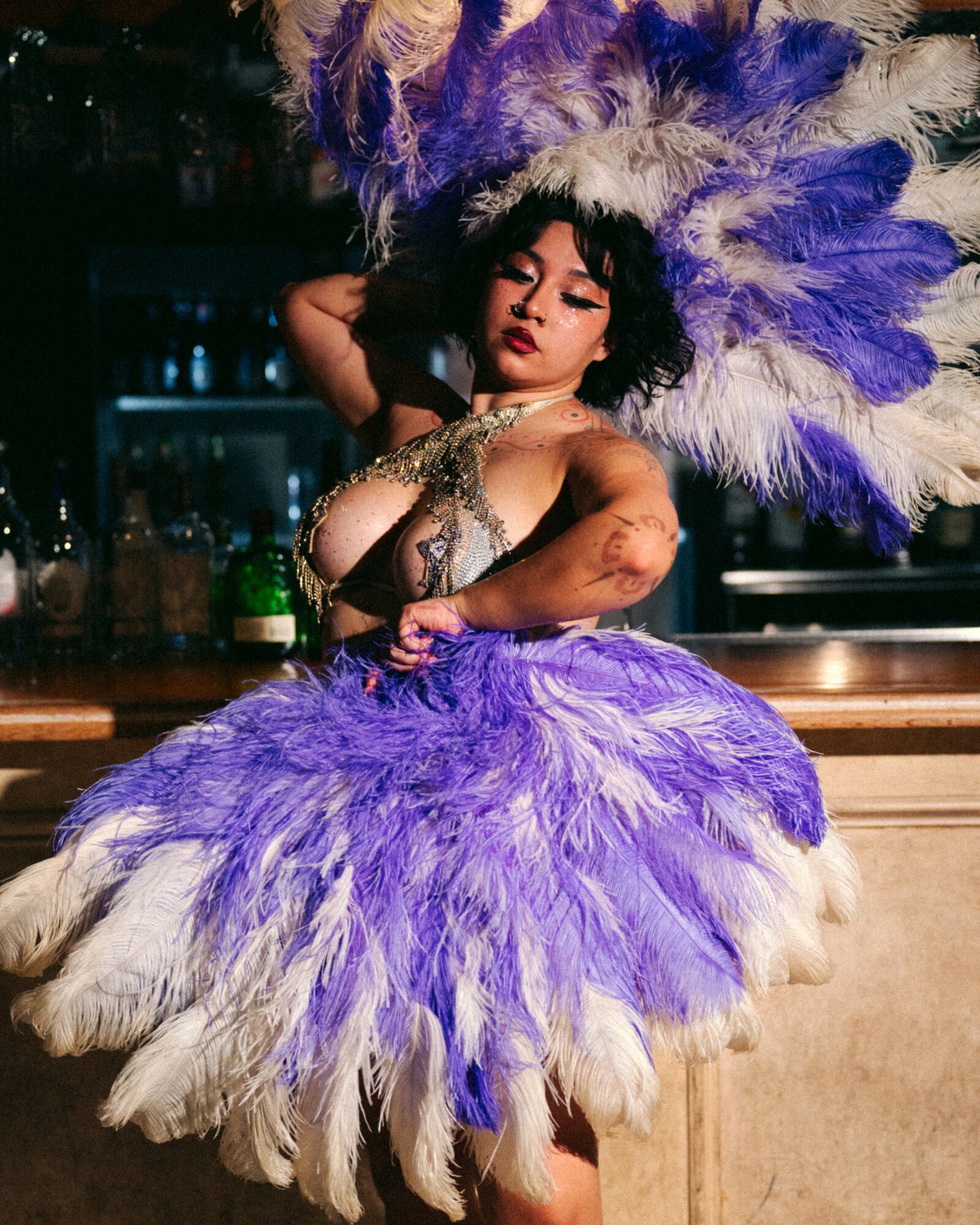 Person in elaborate purple and white feathered costume with headpiece, posing with hand on hip, in a bar setting.
