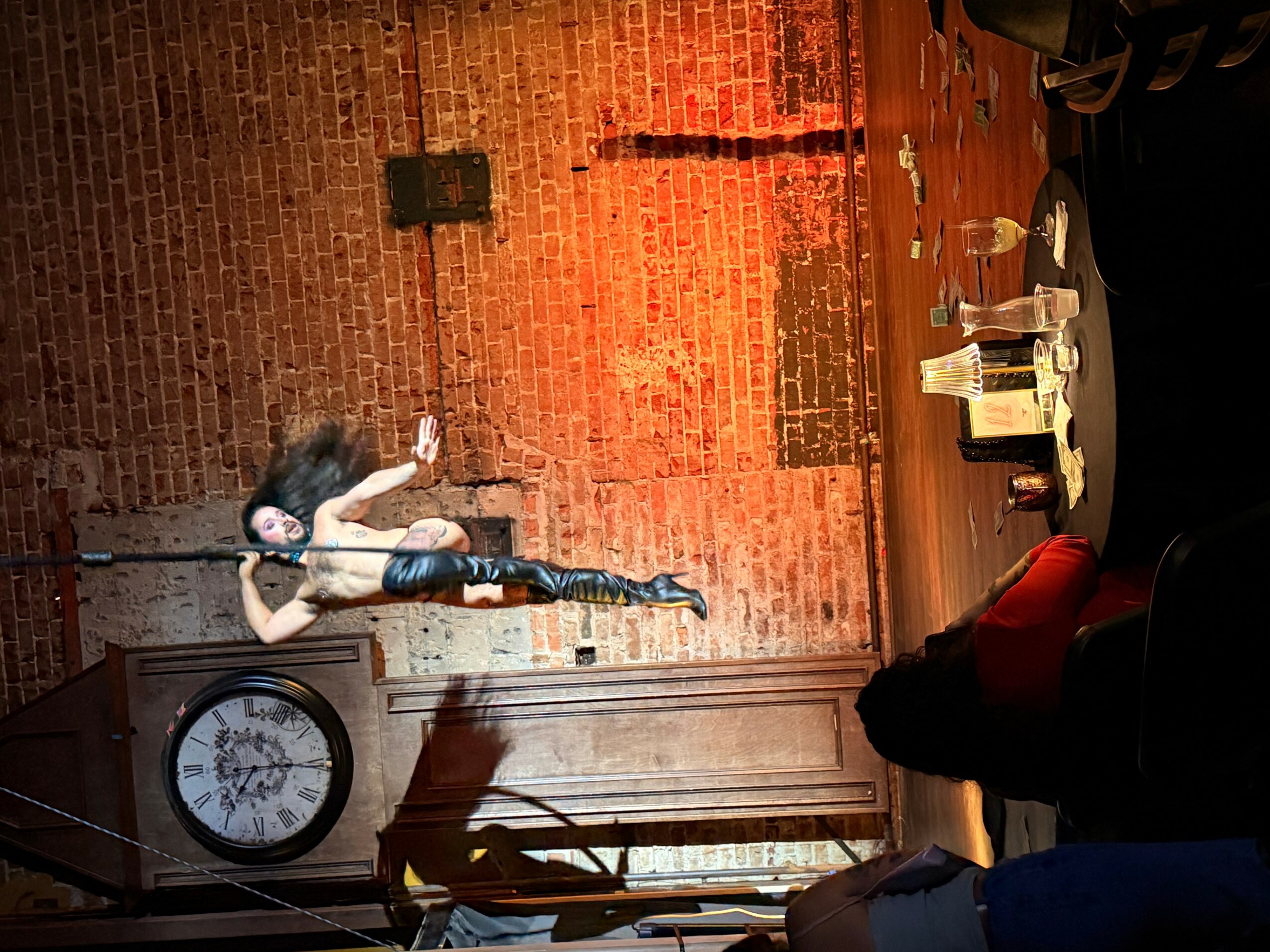 Performer on stage with a microphone, brick wall background, clock, and table with drinks, audience visible.