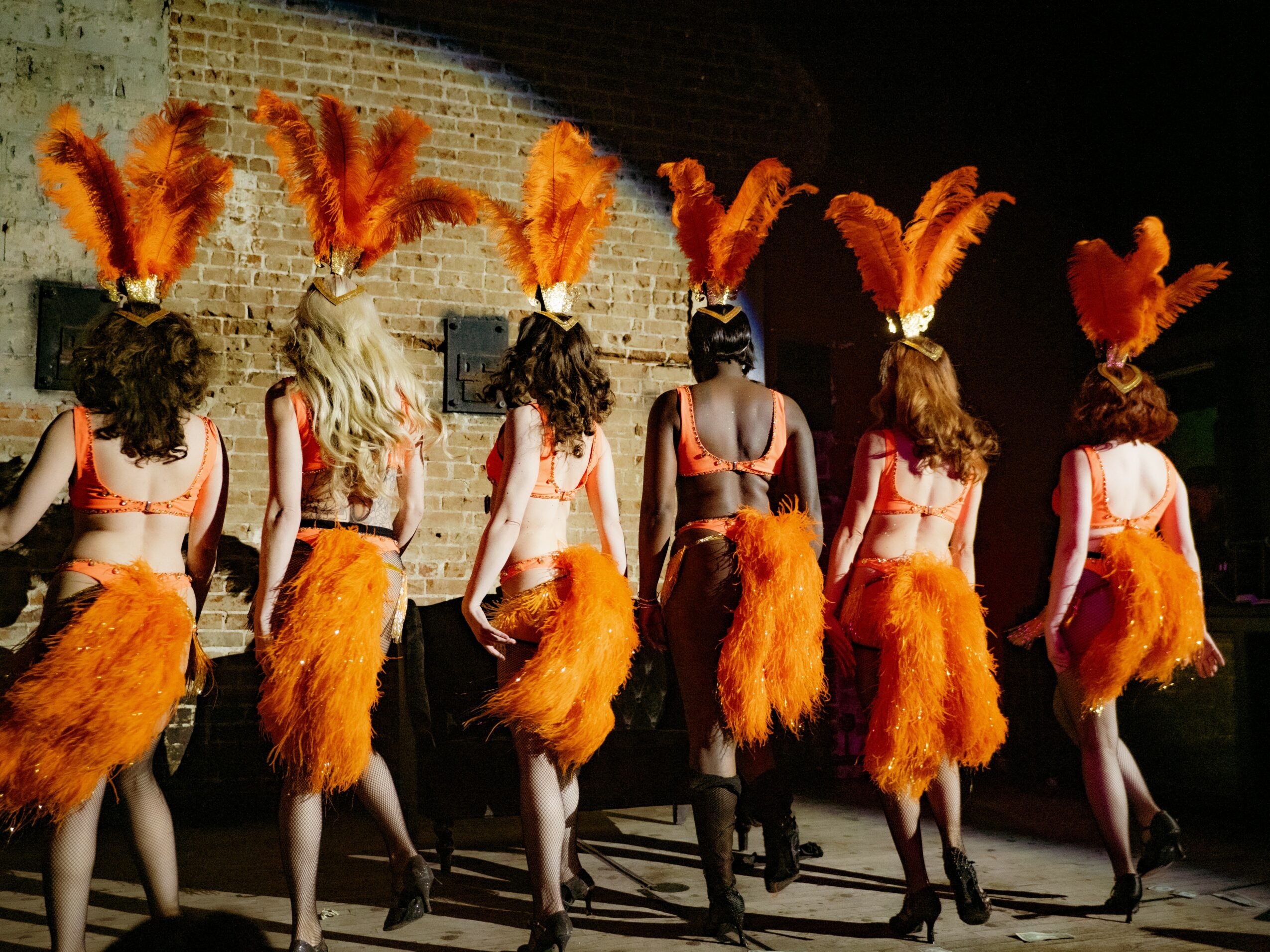 Six performers in costumes with orange feathered headpieces and skirts, facing away, on a stage with a brick wall background.