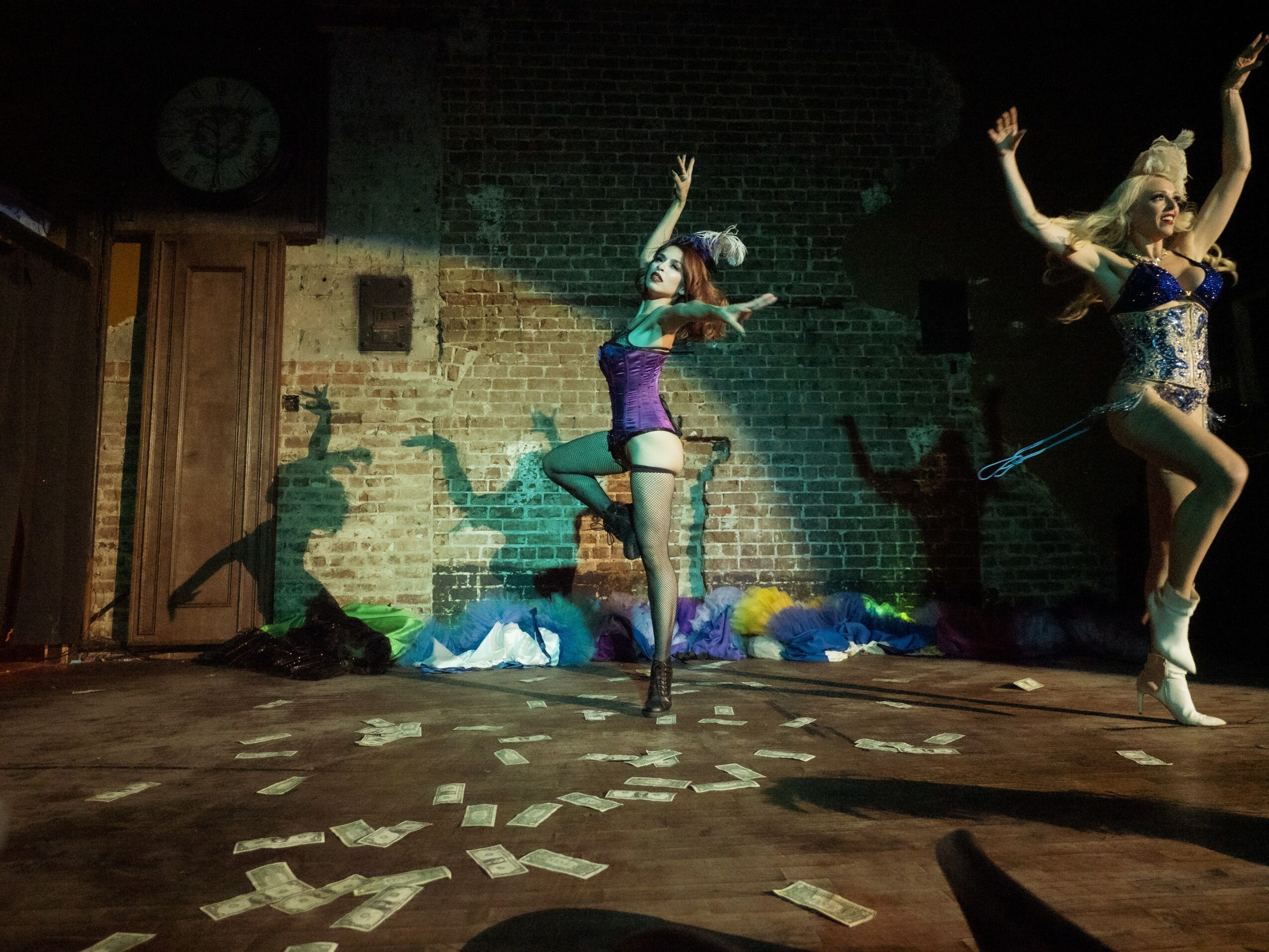Two performers in colorful costumes dance on a stage with scattered papers, brick wall background, and a clock on the wall.