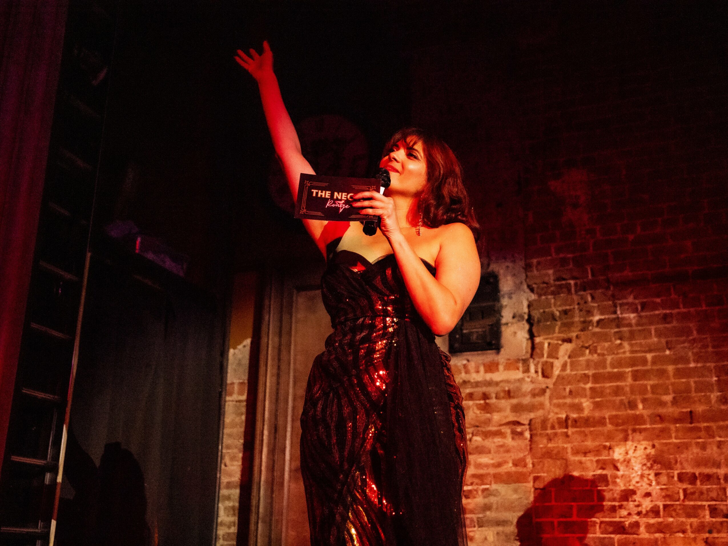 Woman on stage with brick wall background, holding a microphone and raising her hand, wearing a black dress with red accents.