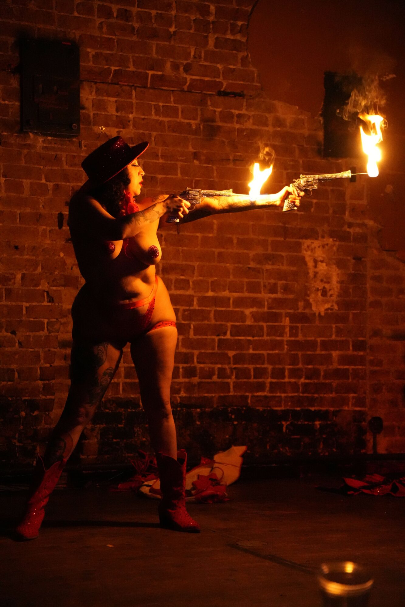 Person performing fire breathing against brick wall at night, flames visible from mouth and torch, dark surroundings.