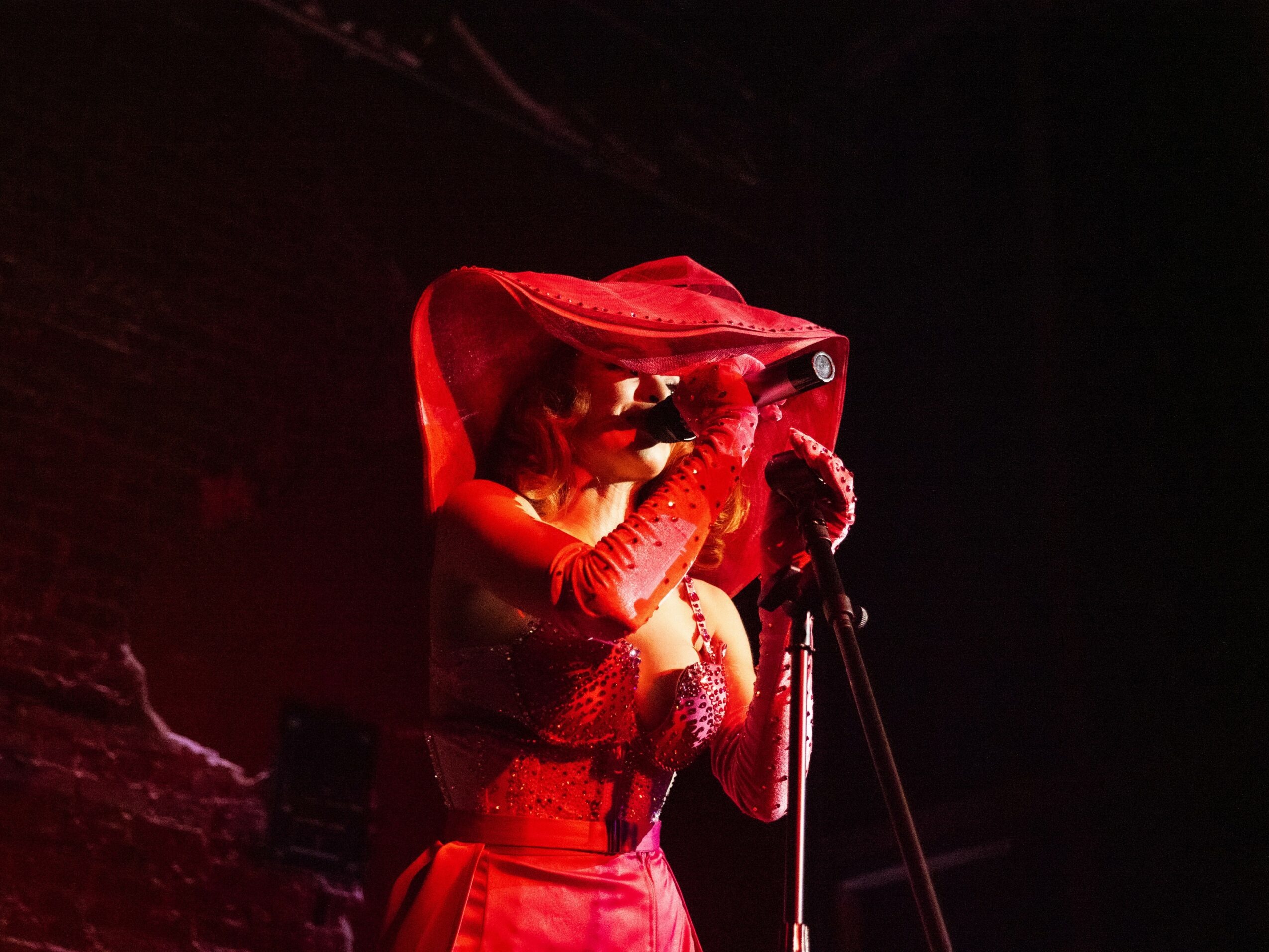 Person performing on stage with a red hooded costume, holding a microphone, dark background.
