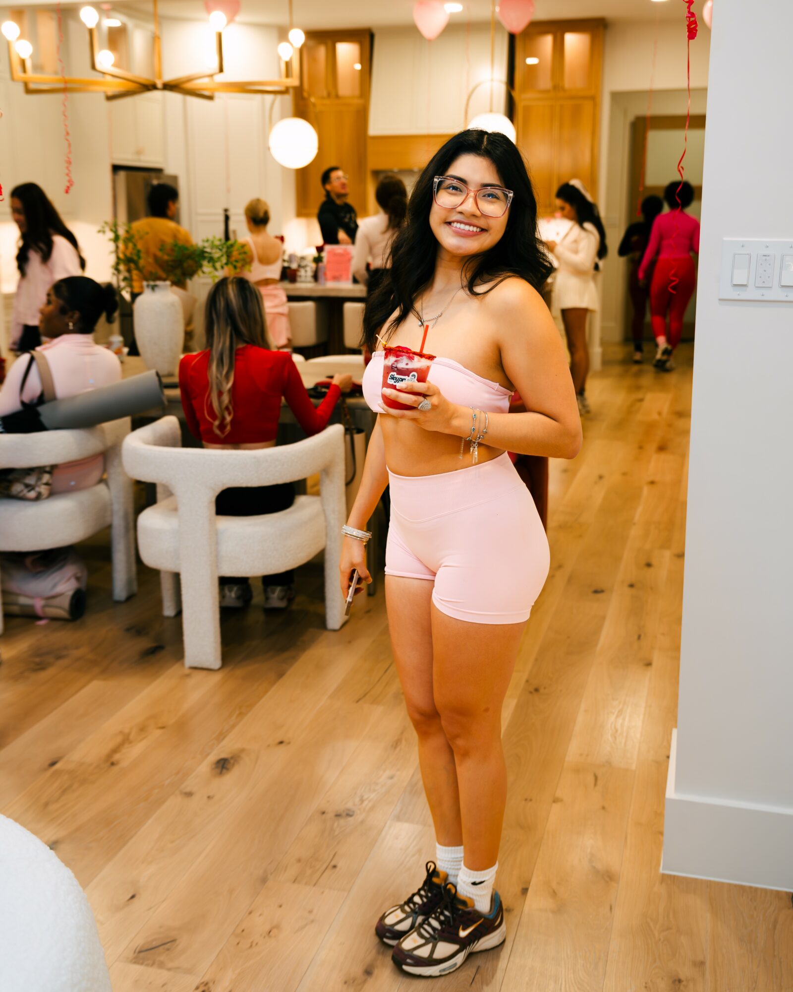 Young woman smiling, wearing glasses, a crop top, shorts, and sneakers, holding a drink at a party with people in background.