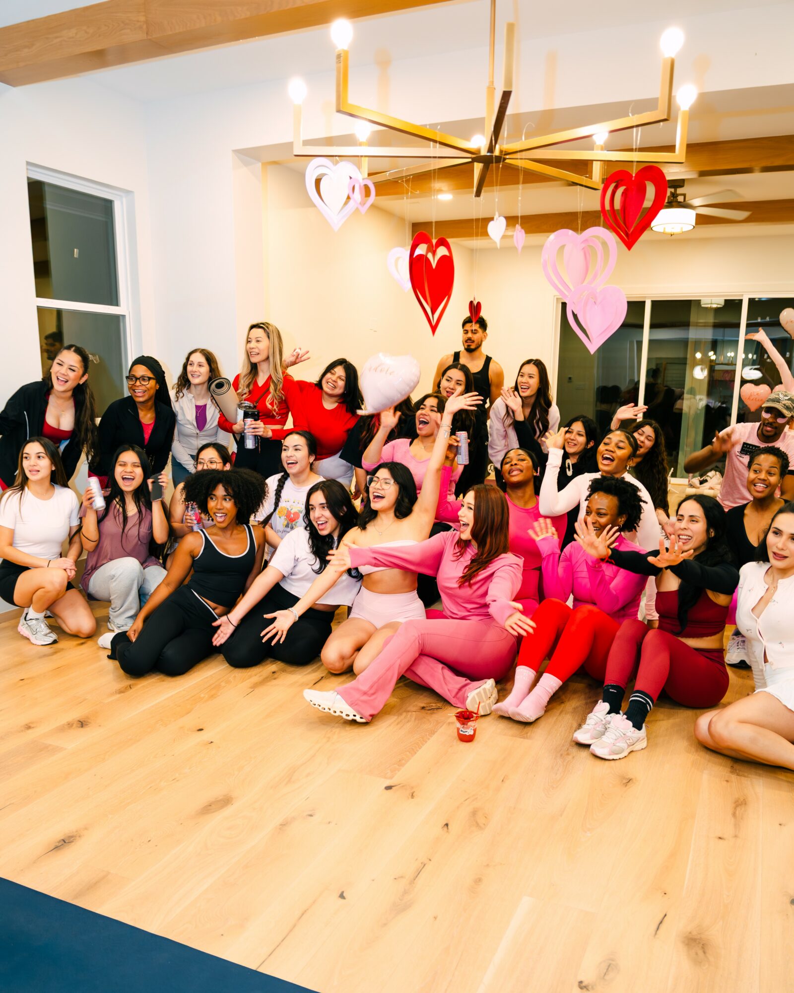 Group of people celebrating indoors with heart-shaped balloons, some sitting and some standing, in a well-lit room with wooden floor.