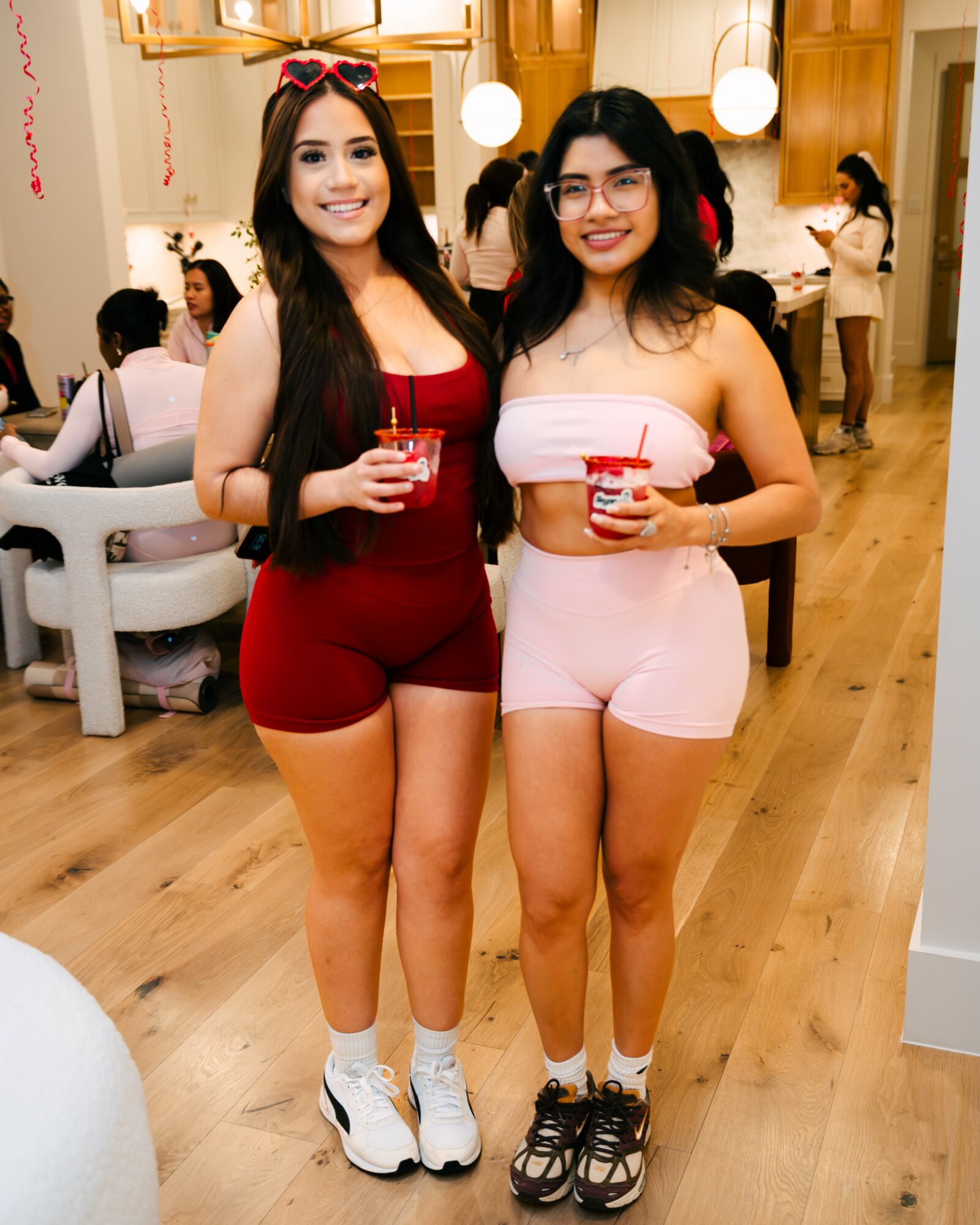 Two women stand together holding drinks in a lively indoor setting with wooden floors and warm lighting.