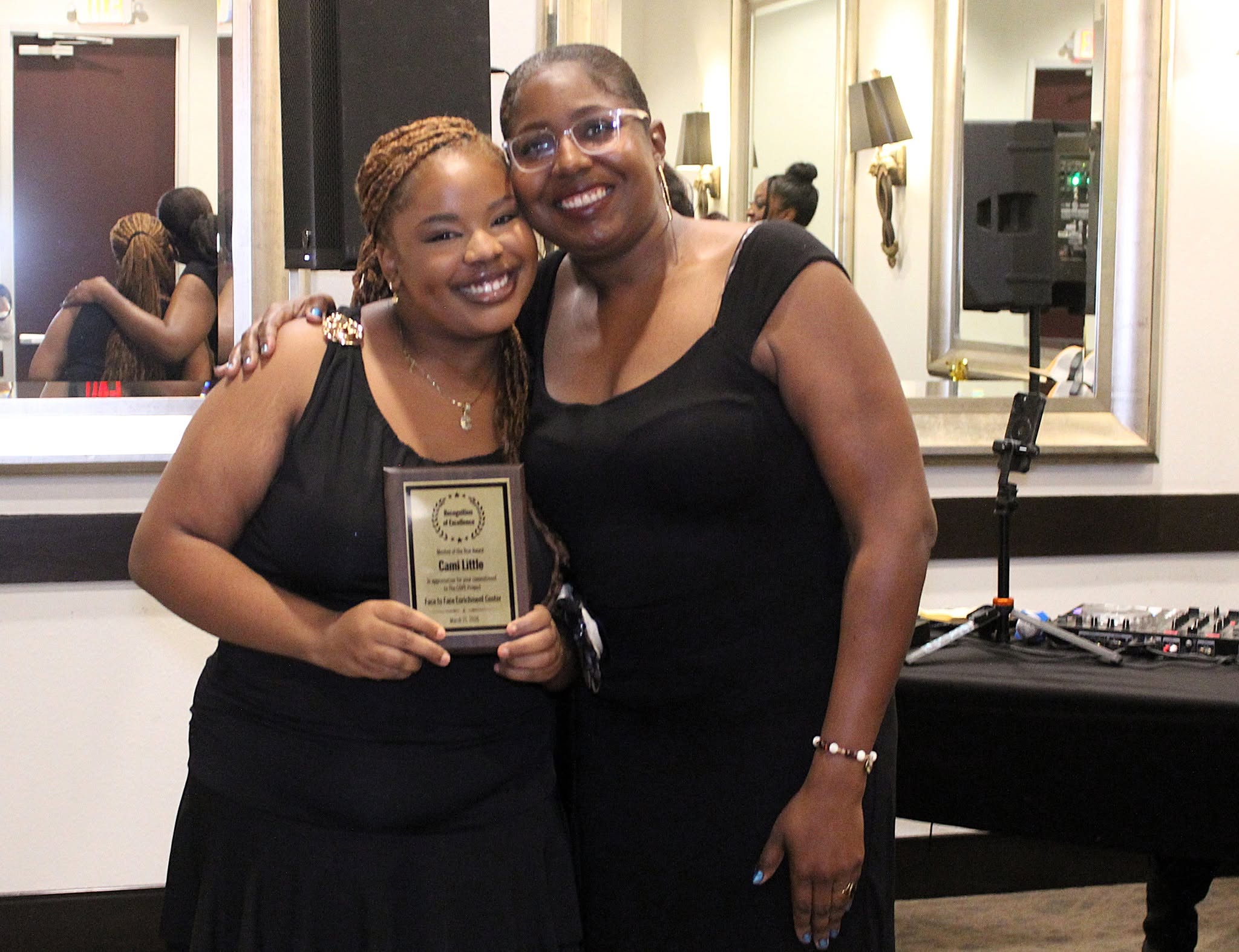 Two women smiling, one holding a plaque, standing in a room with mirrors and a piano in the background.