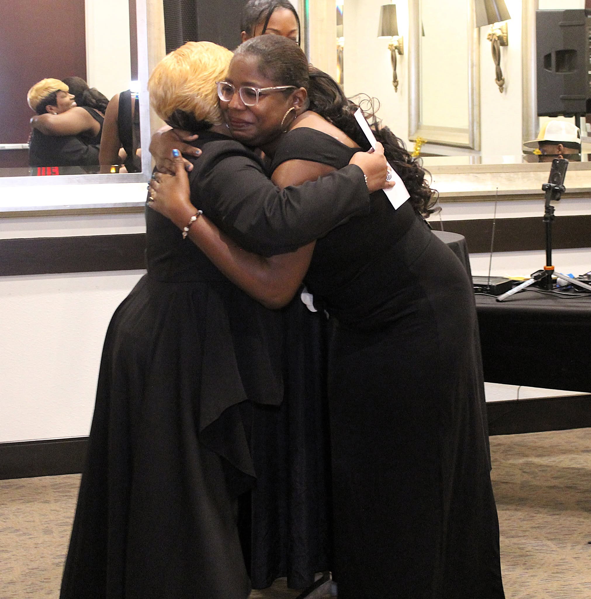 Two women hugging warmly in a room with a mirror and a table, both wearing black dresses.