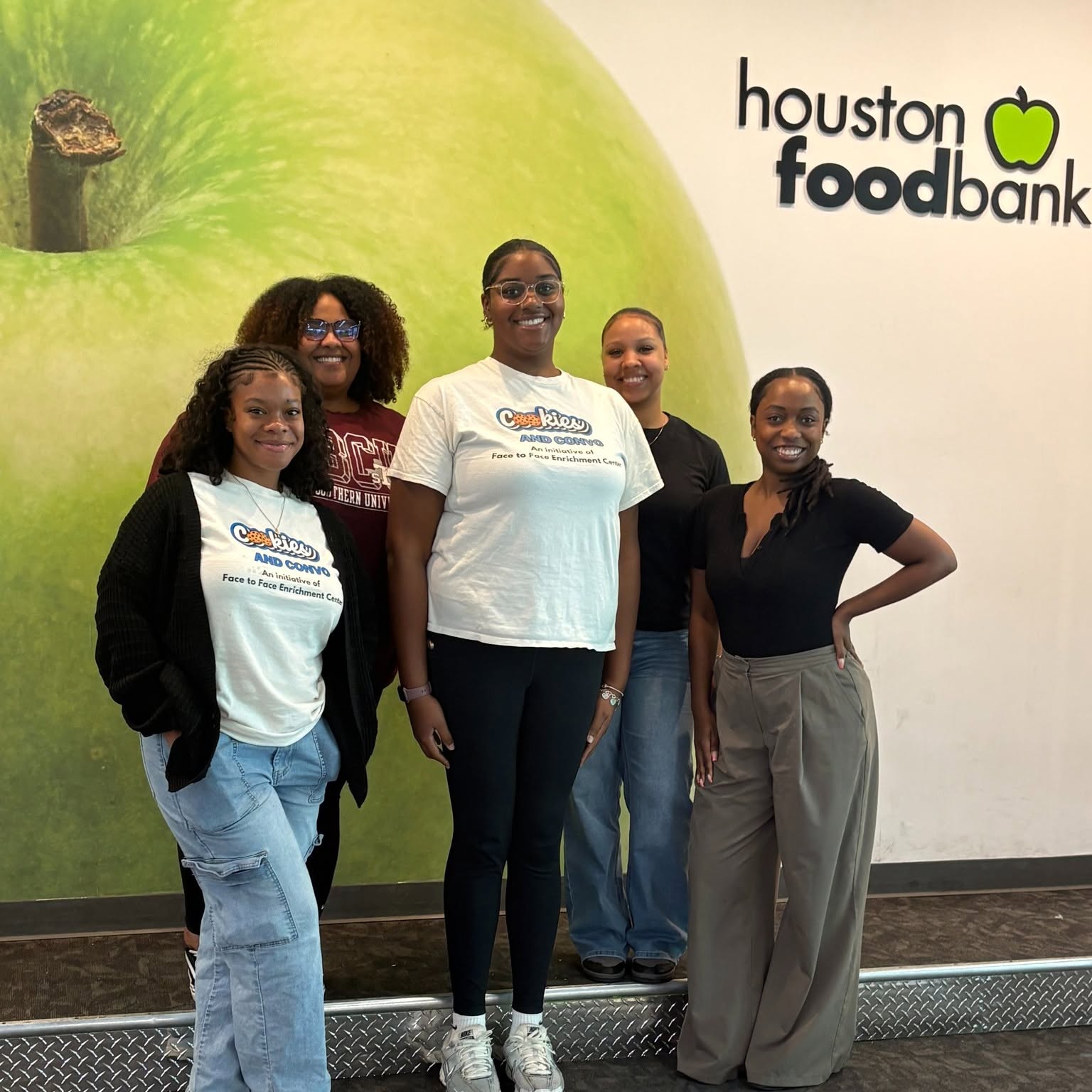 Five women standing together indoors in front of a wall with a green apple and Houston Food Bank logo.