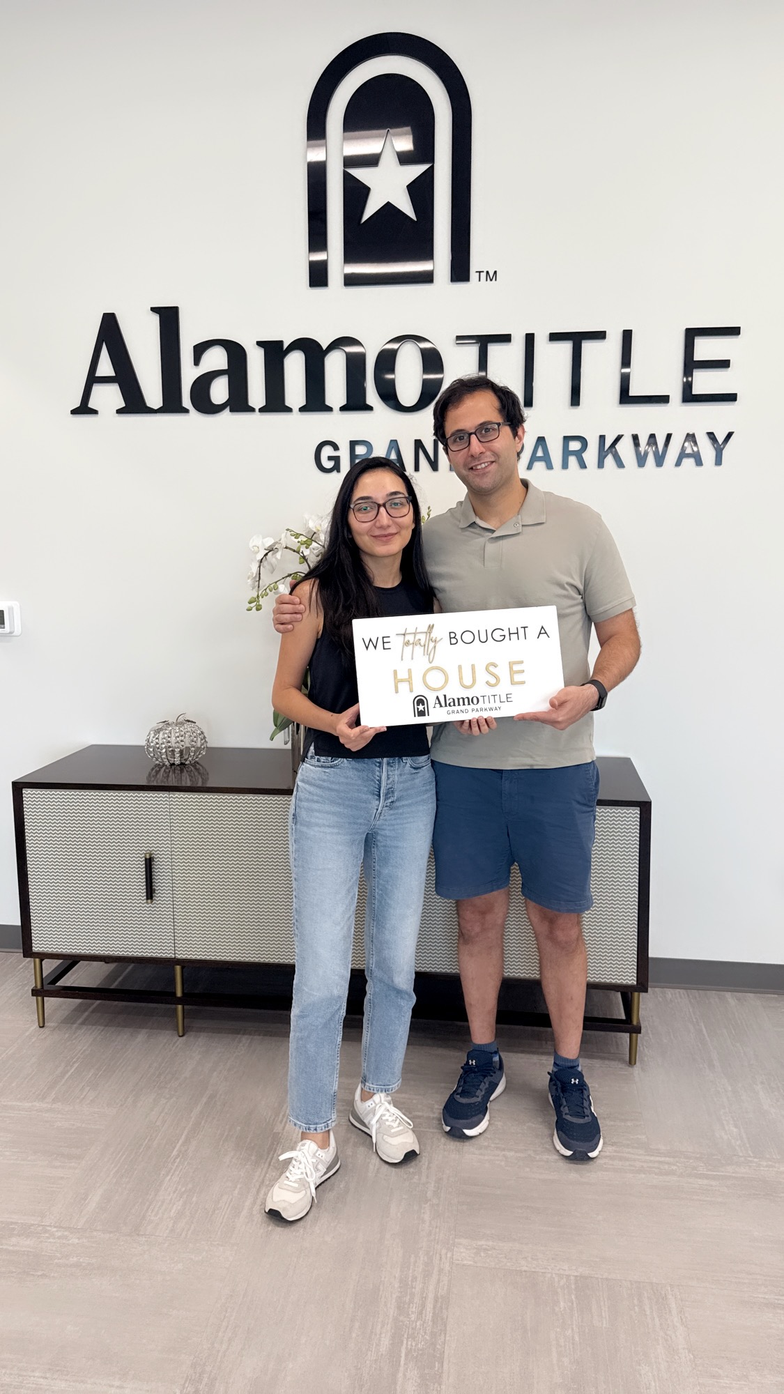 Two people standing in front of a wall with the Alamo Drafthouse logo, holding a sign that says 'We just bought a house.'