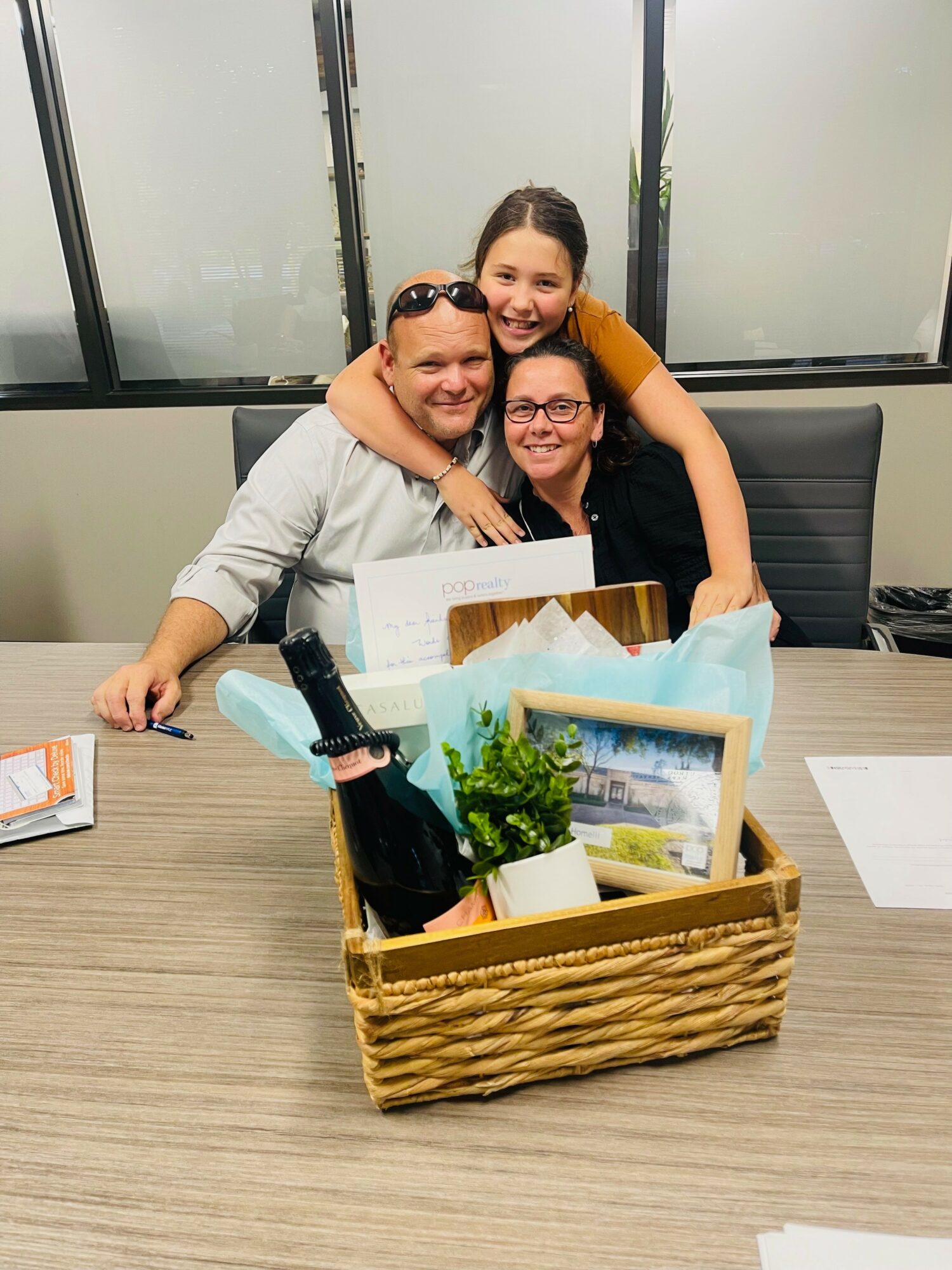 Three people smiling and hugging at a table with a basket of office supplies and framed photos.