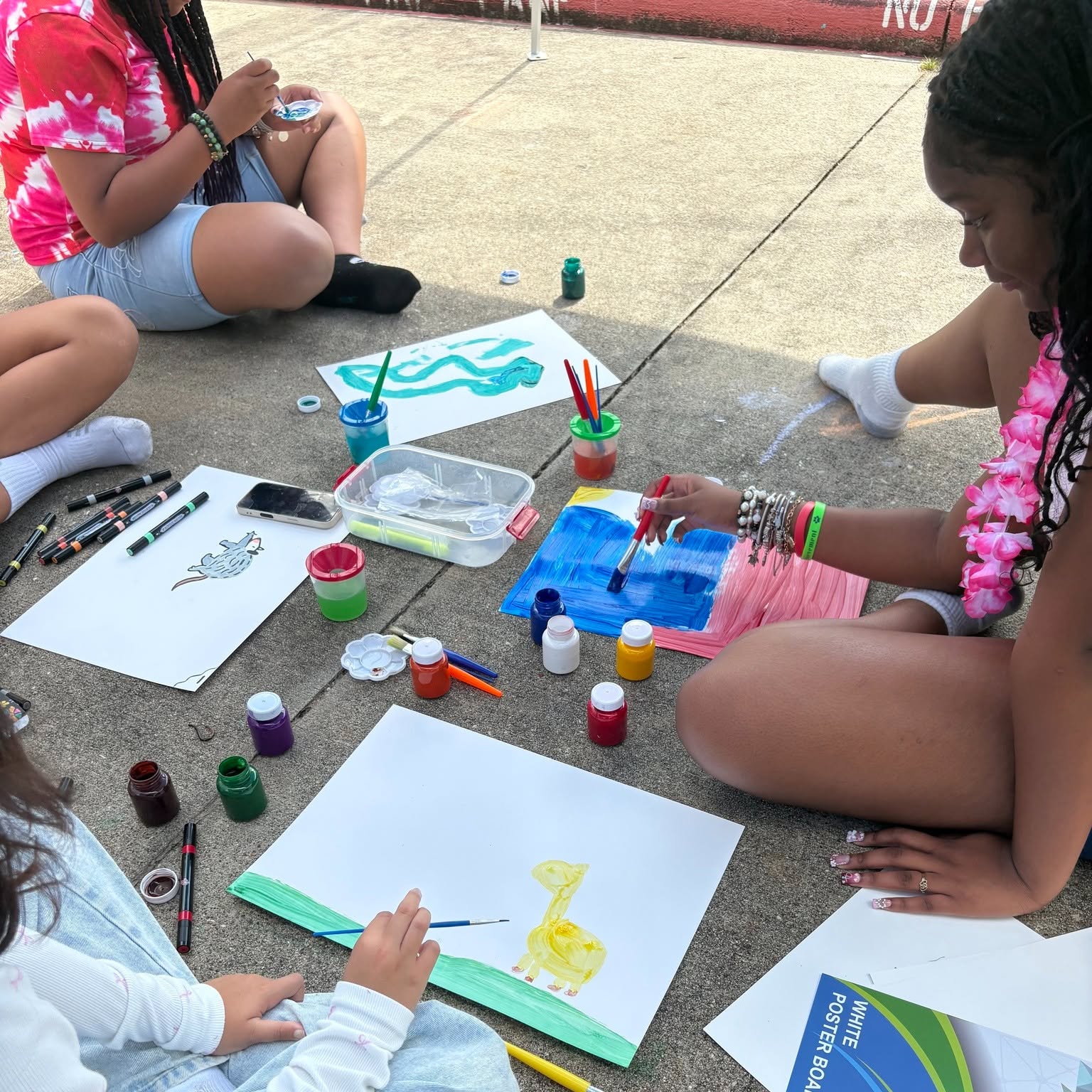 Children sitting on the ground painting on paper with various art supplies around them.