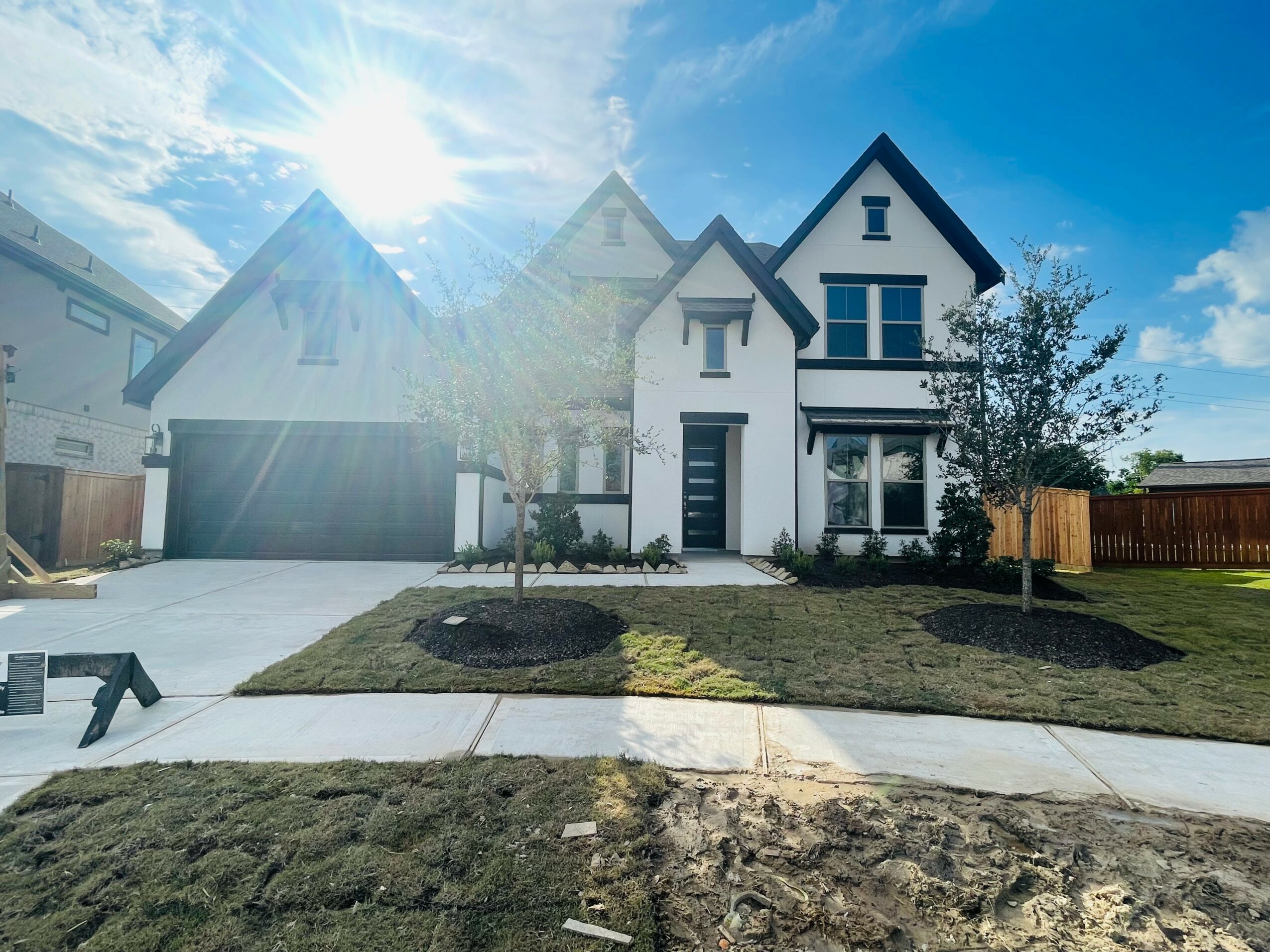 Modern two-story house with a white exterior, black roof, and large windows, surrounded by a lawn and trees, under a sunny sky.
