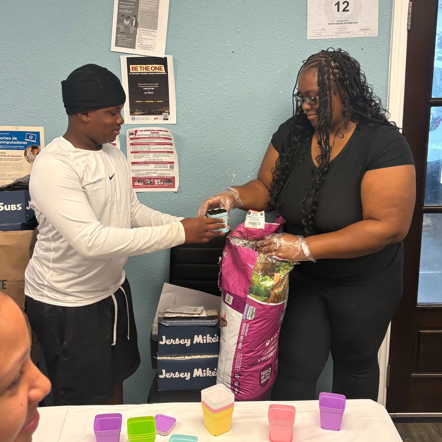 Two women exchanging a small item inside a store, one woman holding a large bag of pet food, table with colorful cups in foreground.