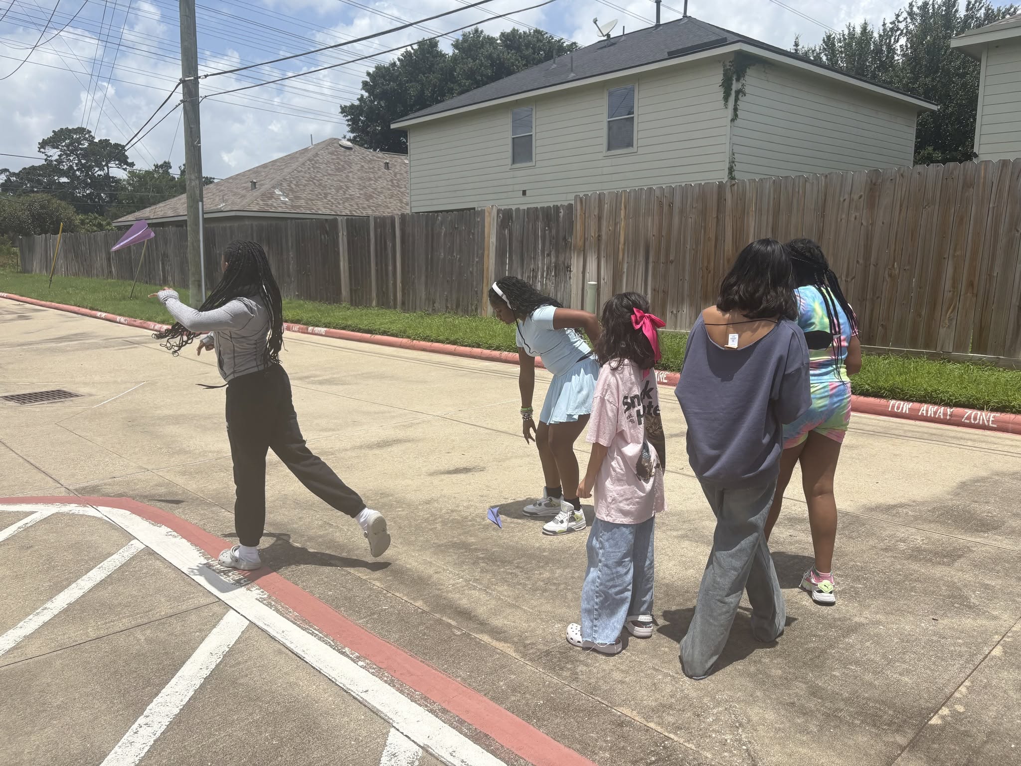 Group of children standing on sidewalk near a fence, some looking at a person walking away, in a residential area.