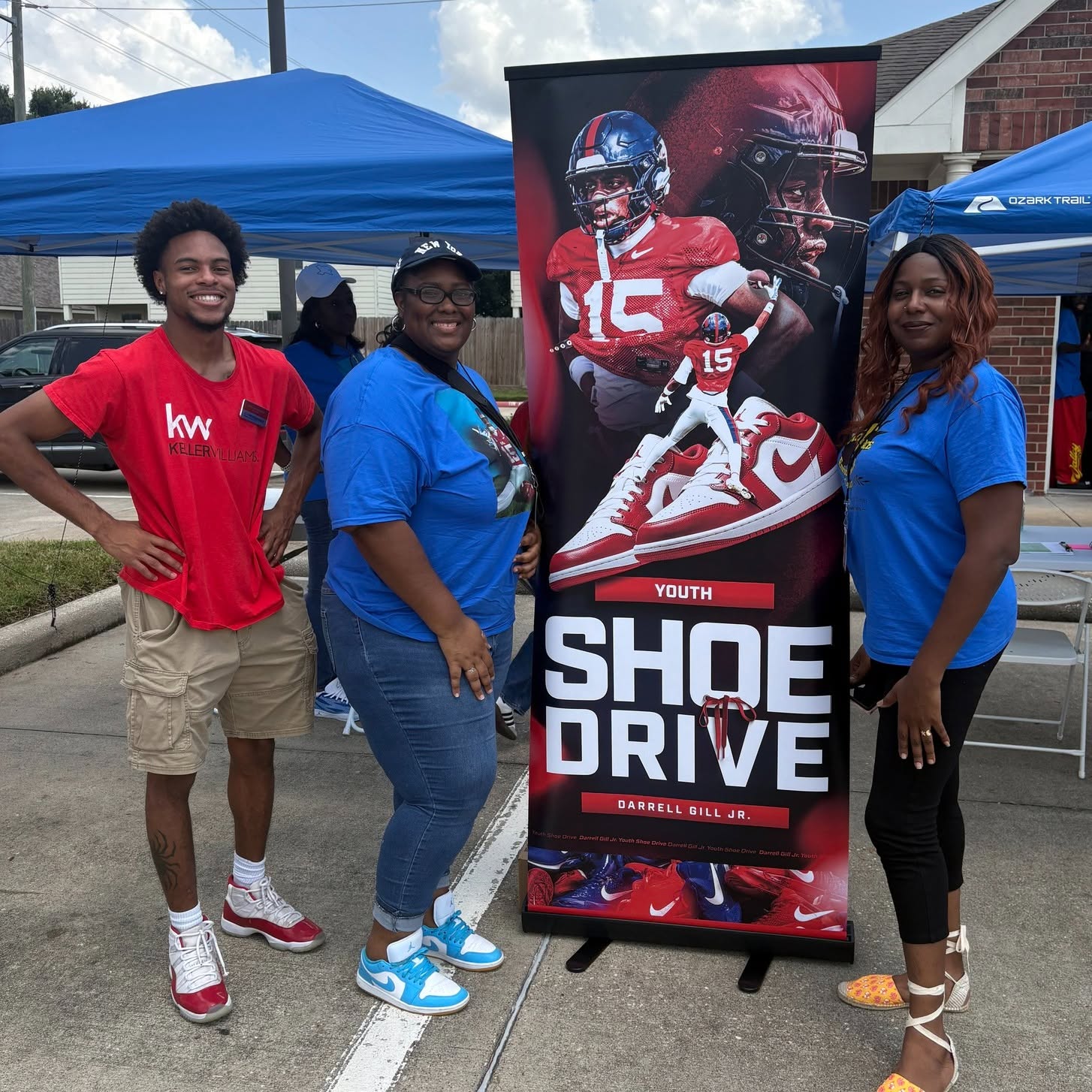Three people standing outdoors next to a banner for a youth shoe drive, with tents in the background.