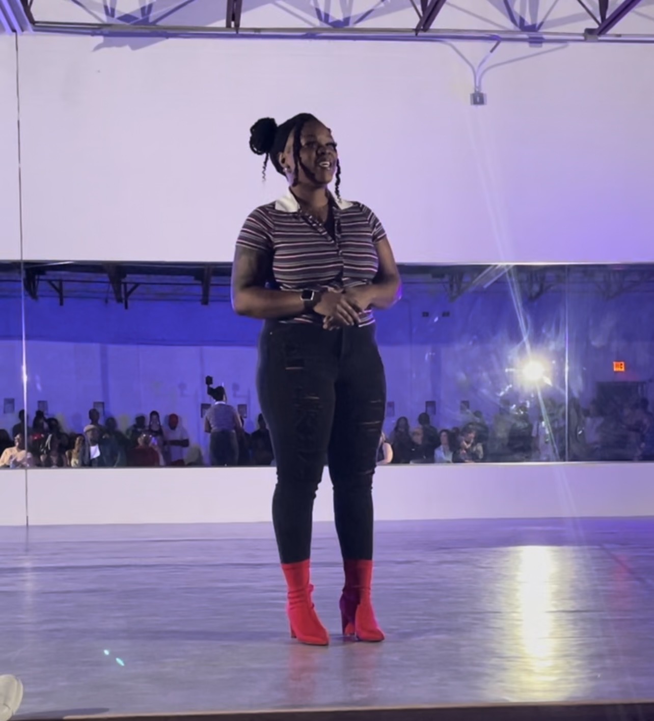 Woman standing on stage with a large mirror behind her, wearing striped shirt, black pants, and red boots, in a dance studio.