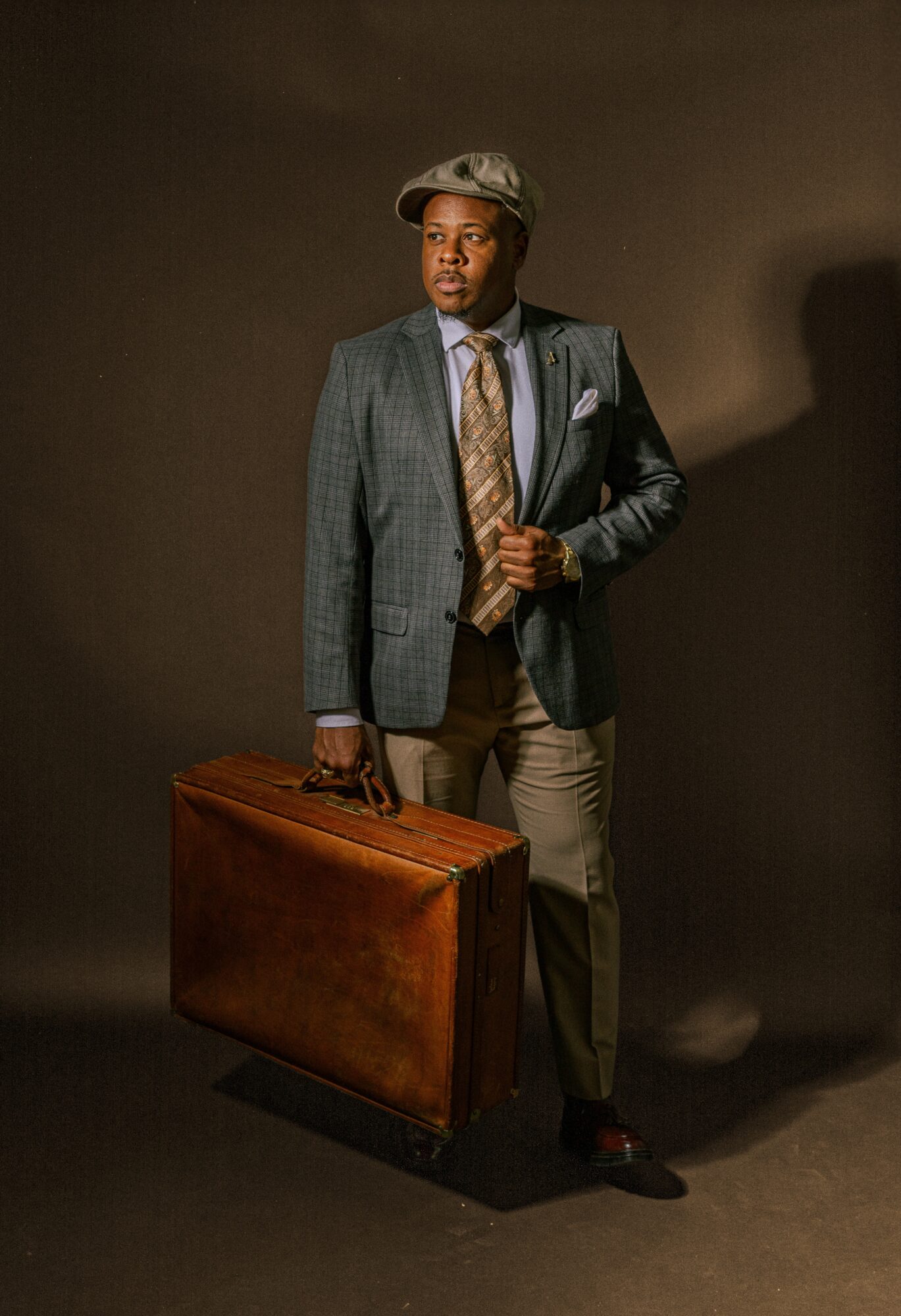 Man in suit holding a briefcase, wearing a hat, standing against a dark background.